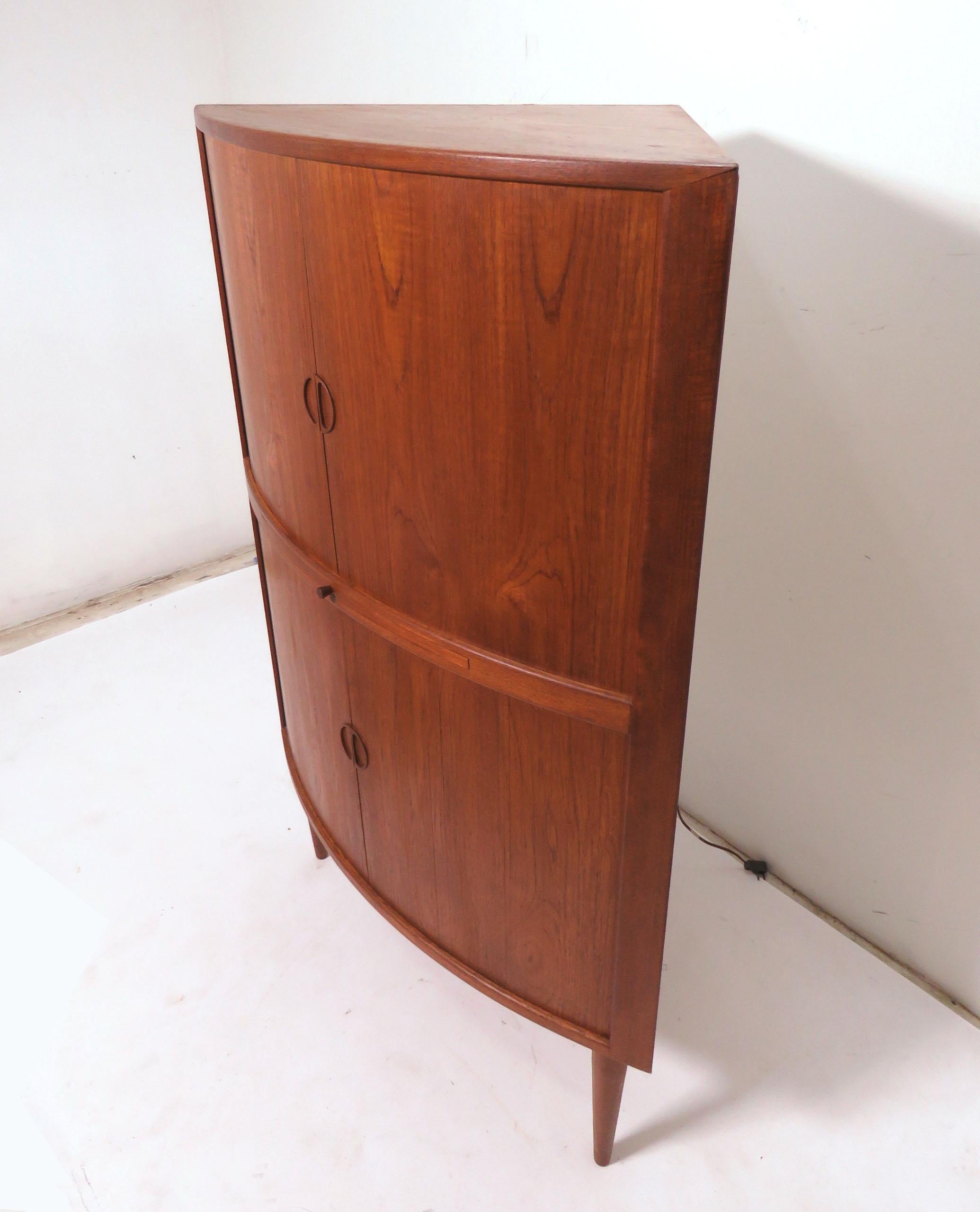 Danish teak corner cabinet with tambour doors. Upper section includes a backlight, mirrors, a glass shelf, and two storage drawers. pull-out shelf for preparing drinks. Lower section for storage of bottles.