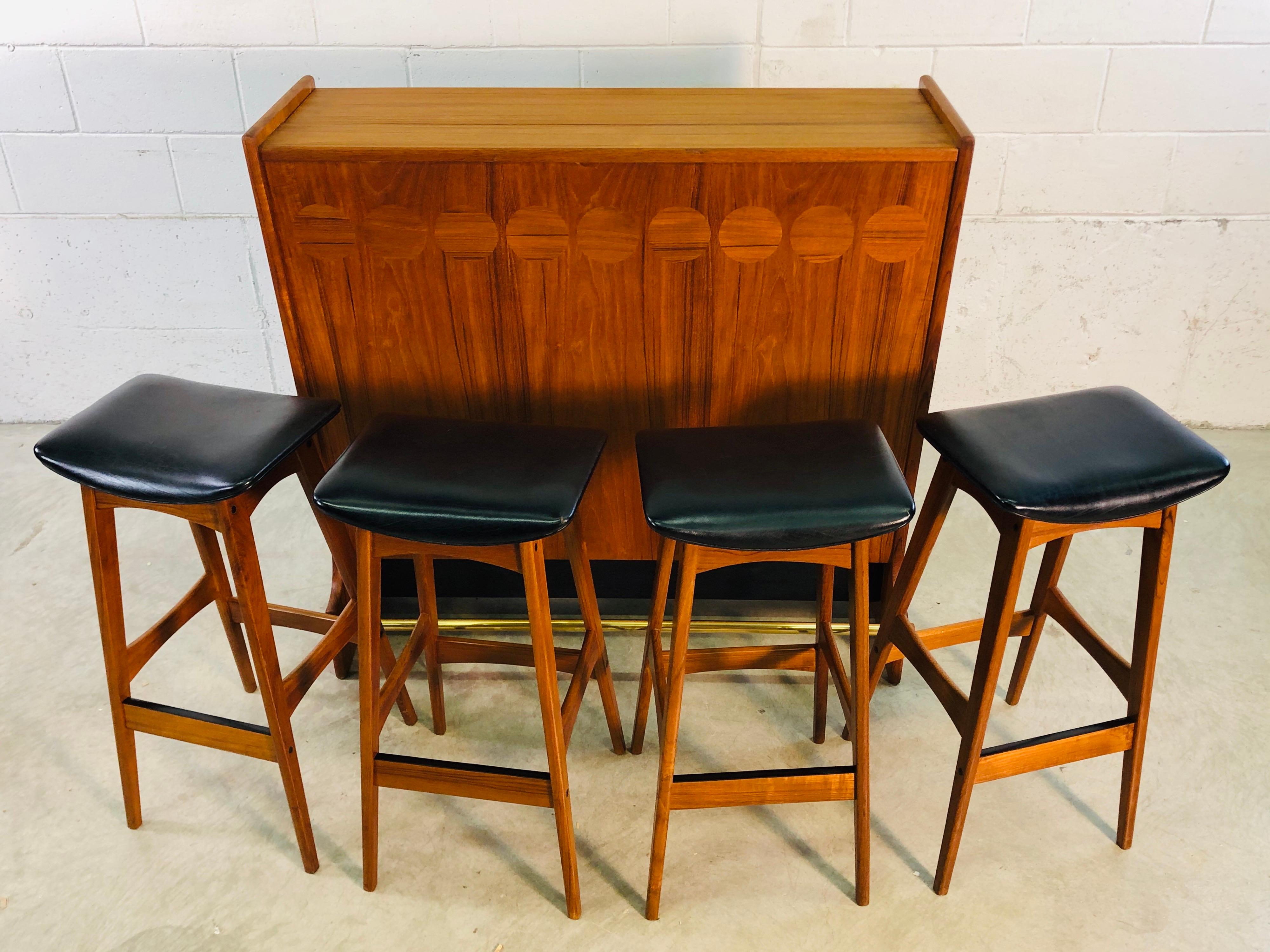 Vintage Danish teak dry bar and four stools designed by Johannes Anderson for BRDR Anderson Vejen. The teak dry bar holds a total of 18 bottles, comes with glass shelves and a mirror inside. The bar is accented with a brass footrest and black