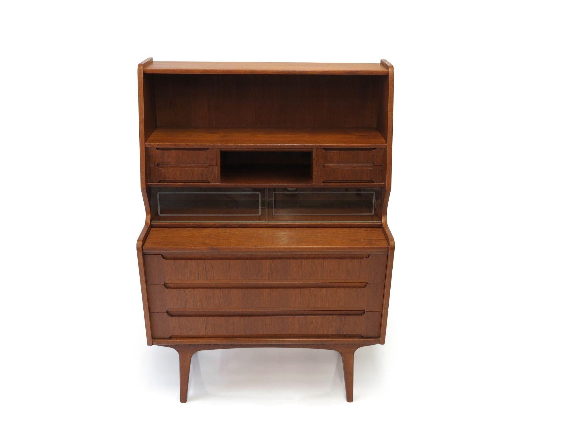 Danish teak secretary desk crafted of teak with book-matched grain across the drawers. The secretary features a pull-out writing surface over three drawers. A pair of sliding glass cabinet doors, a hidden vanity mirror, and four small drawers above.
