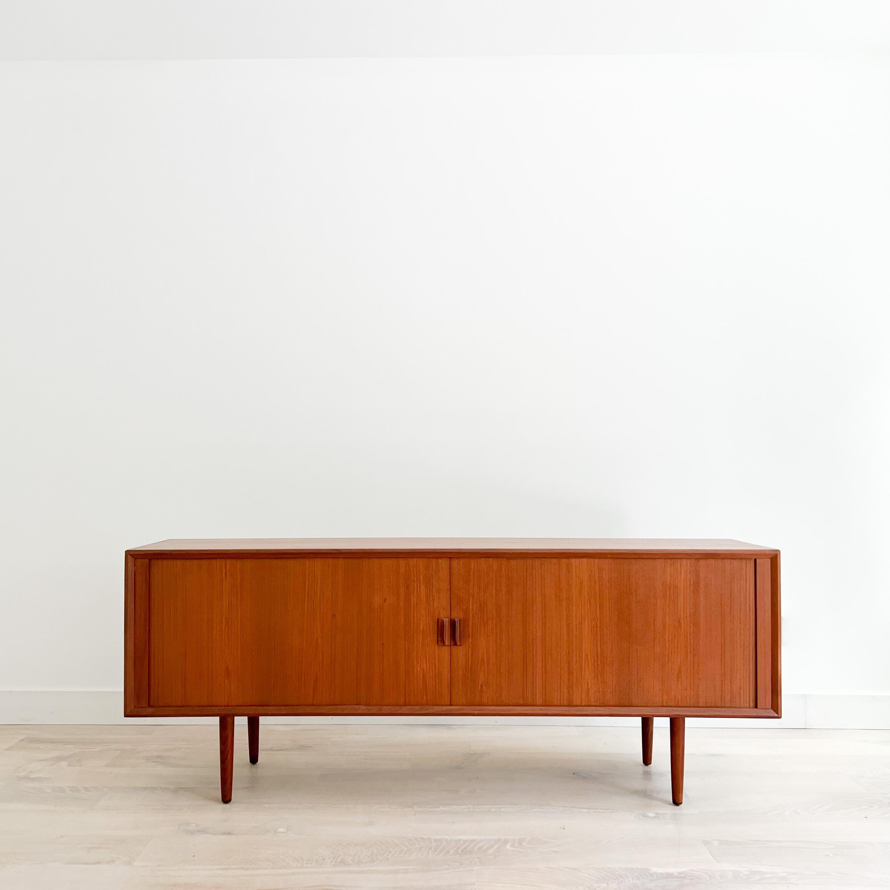 Mid-Century Modern long danish teak credenza with tambour doors on tapered legs. Designed by Svend Aage Larsen. Some light scuffing/scratching from age appropriate wear. The tambour doors open and close with ease. Very slight raised area in the