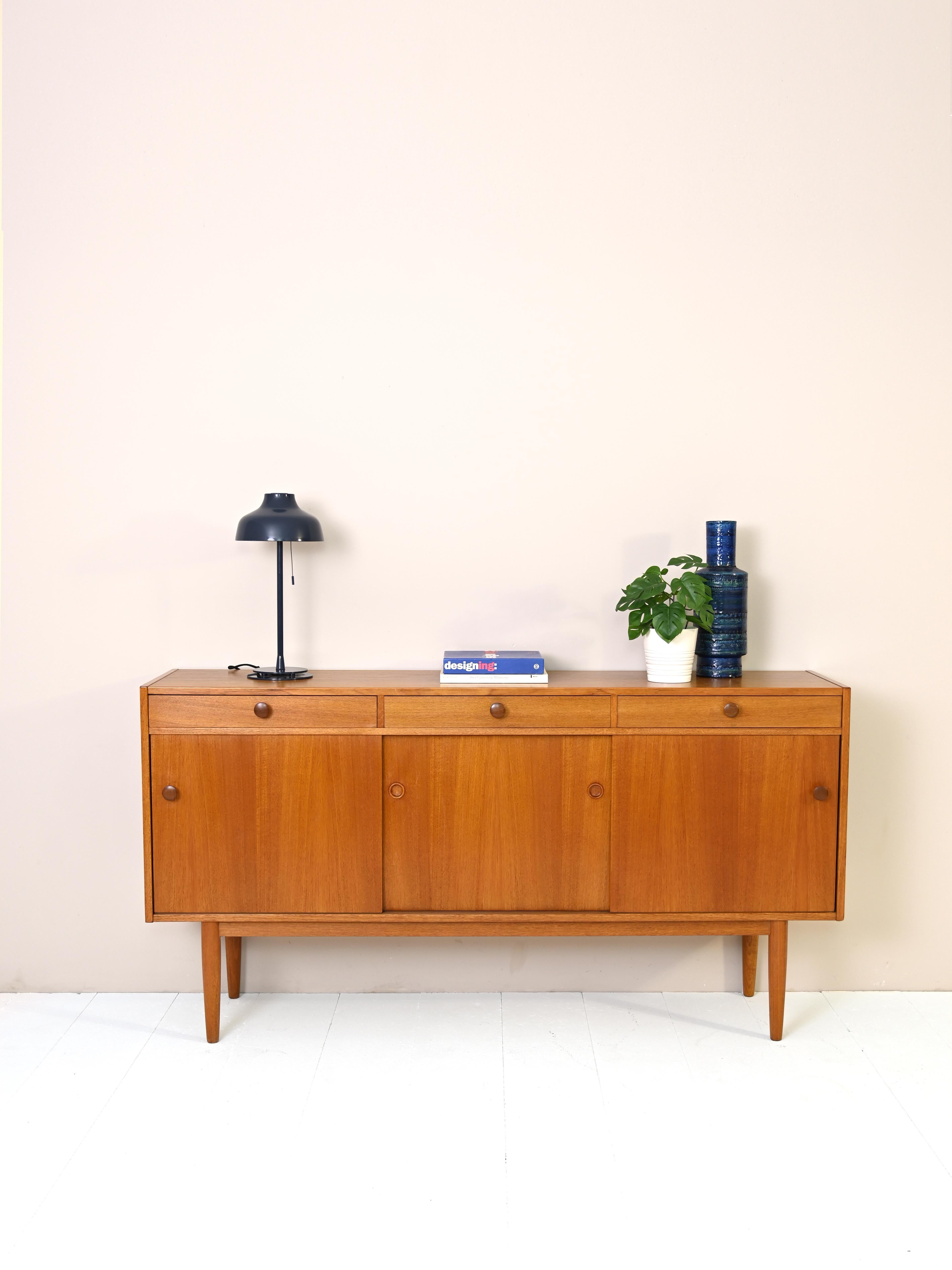 Scandinavian sideboard with stamp of authenticity.
The cabinet features three drawers with wooden knobs and three sliding doors. Inside are three
compartments equipped with a shelf respectively.
The long conical legs give lightness to the