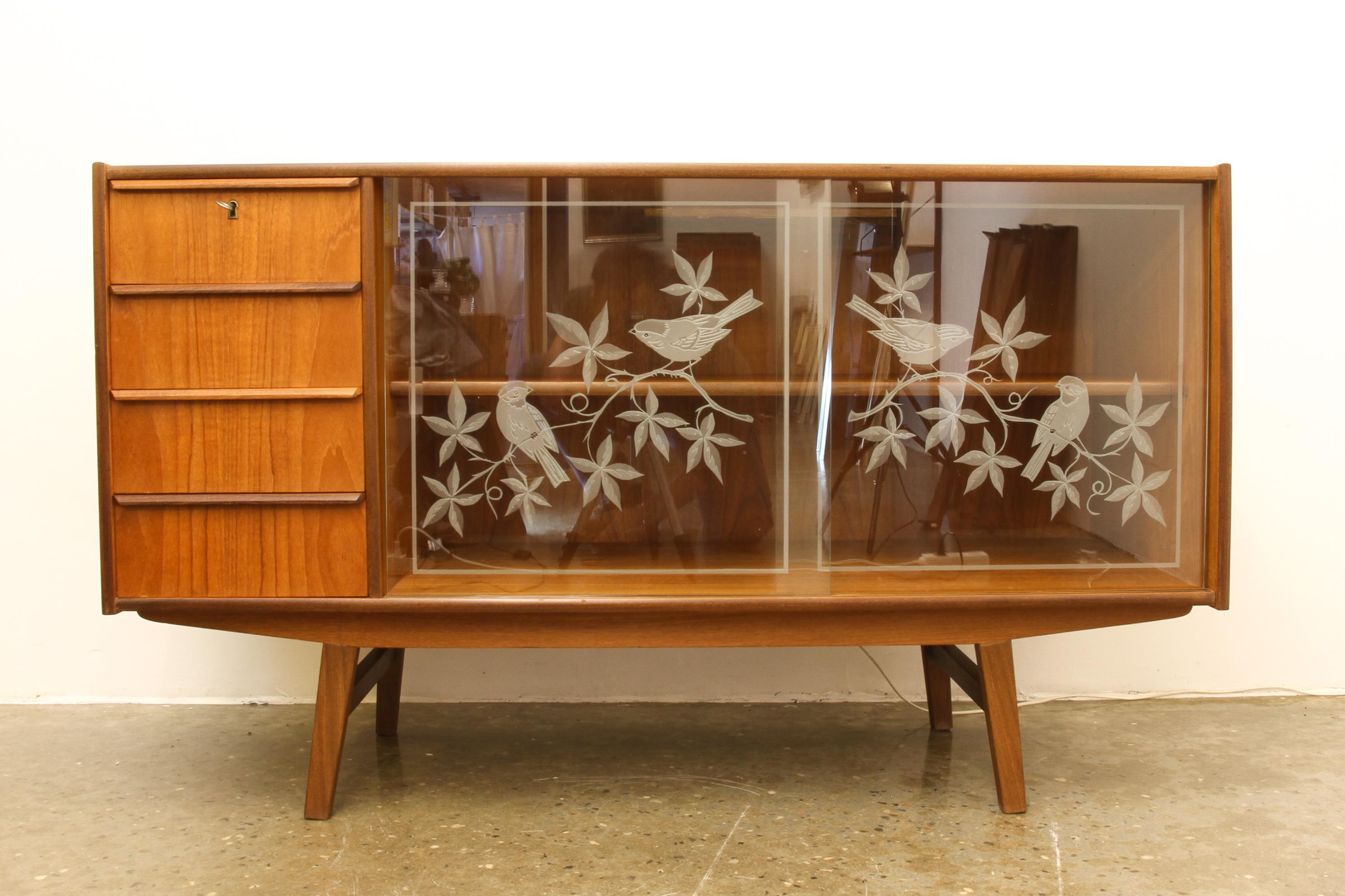 Danish teak sideboard with glass doors 1960s.
Beautiful Danish Mid-Century Modern sideboard with four drawers and compartment with one shelf. Double glass doors with etched bird motif. Built in light behind the glass doors. Standing on four