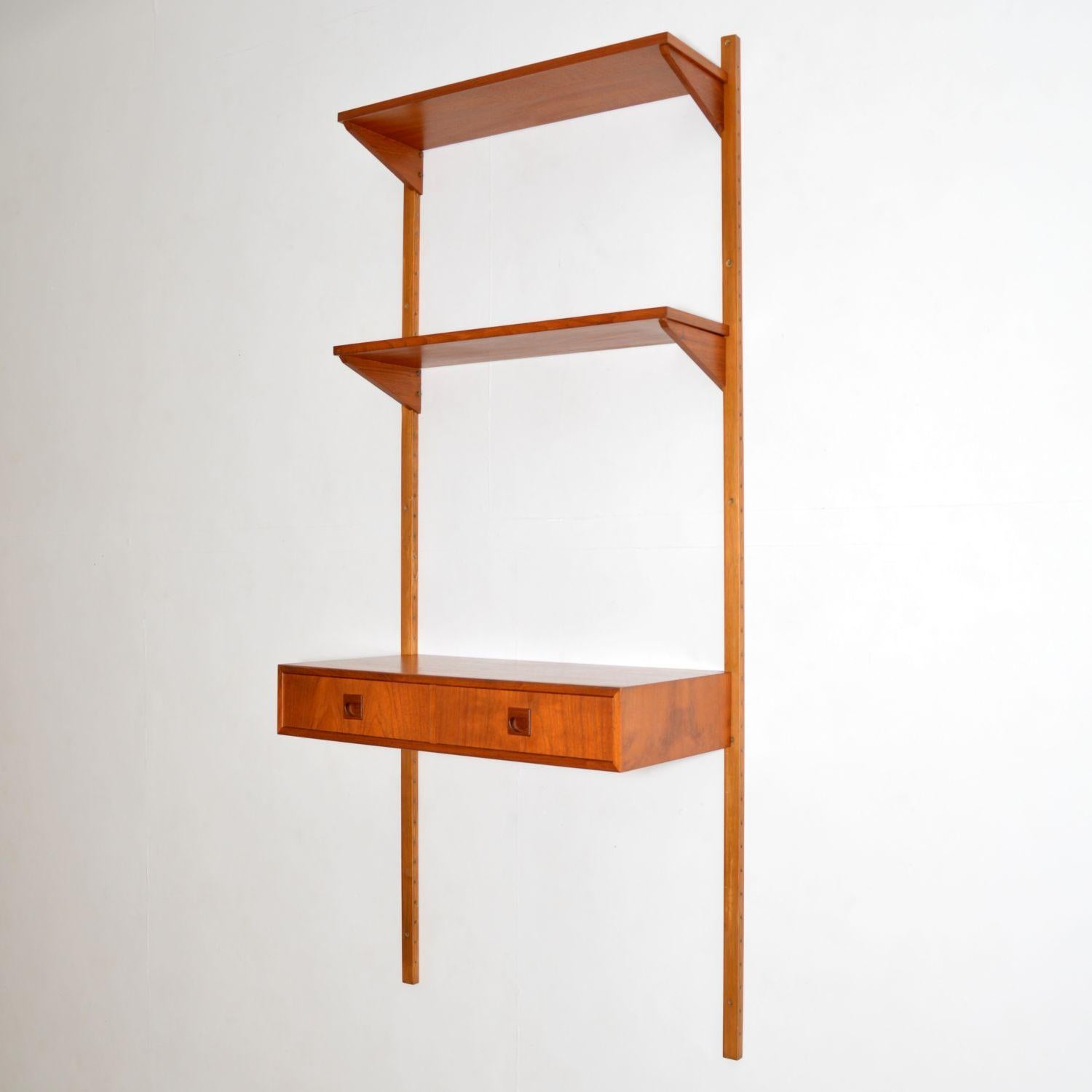 A stylish and versatile teak wall mounting cabinet with desk. This is the PS system, made in Denmark in the 1960s.

This single bay consists of a two drawer desk section, two chevron supported shelves, and two long wall mounting rails.

This is
