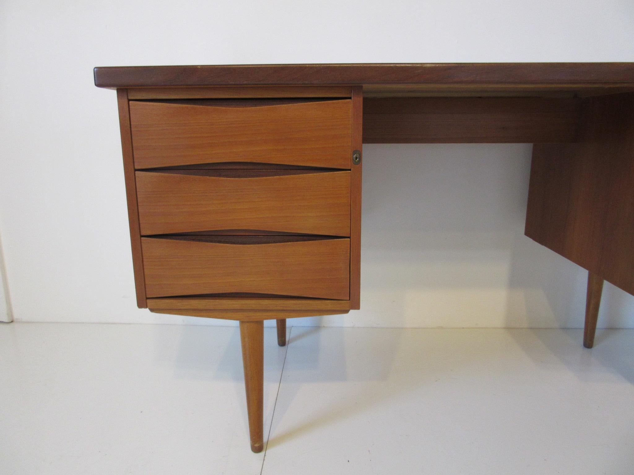 A teak wood desk with conical legs and two banks of drawers one side having a file and the other side three separate drawers with cutout eye pull openings in the style of Vodder. Each bank of drawers lock and includes the original key, made in