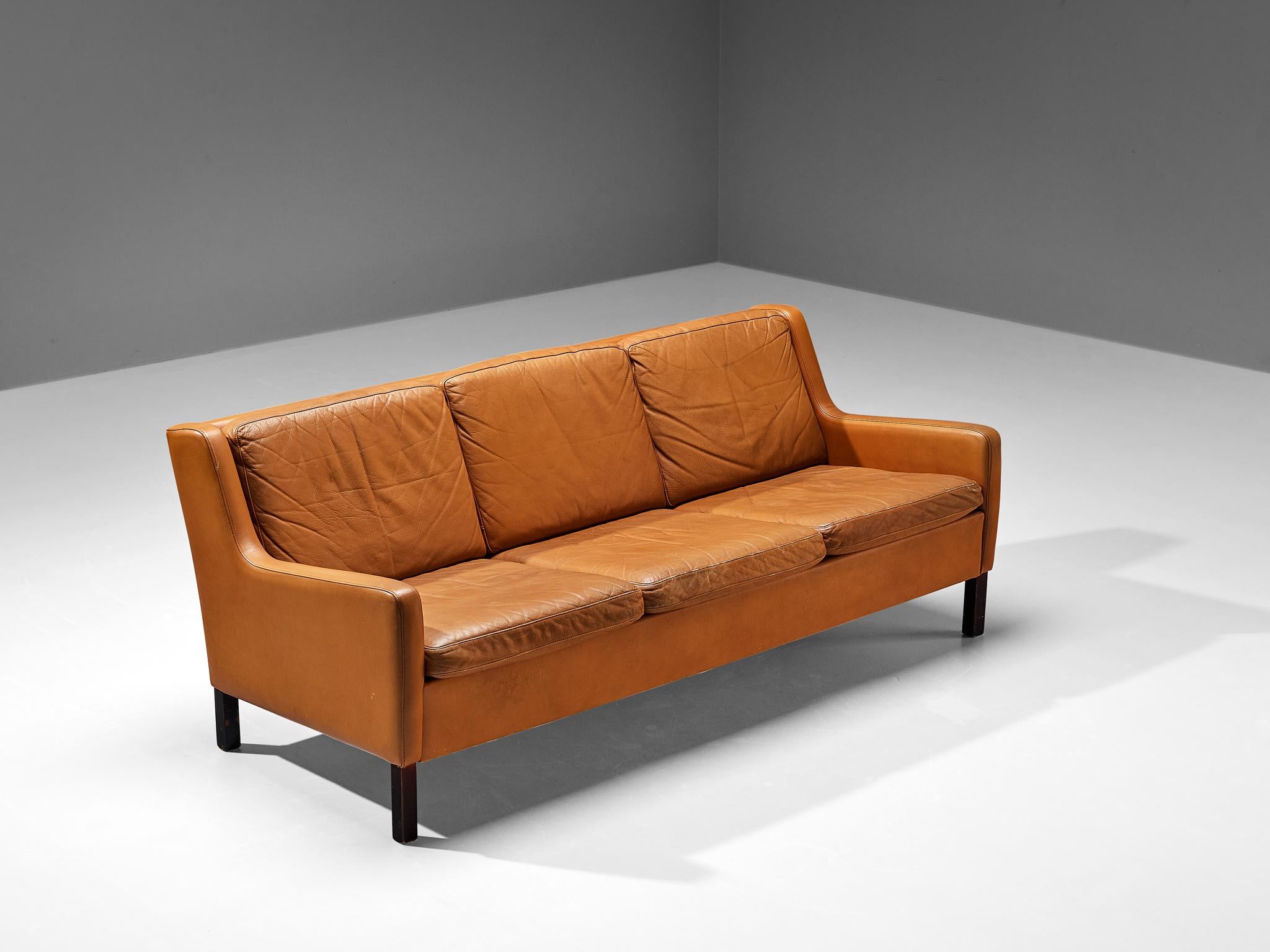 Three-seat sofa, cognac to orange leather and oak, Denmark, ca. 1960. 

This modest and comfortable three-seat sofa that shows traits of the design of Borge Møgensen. The colour palette of the orange-cognac leather and the dark stained legs create a
