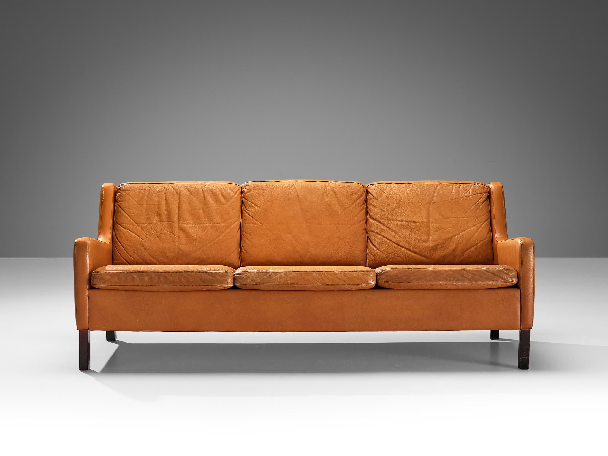 Three-seat sofa, cognac to orange leather and oak, Denmark, ca. 1960. 

This modest and comfortable three-seat sofa that shows traits of the design of Borge Møgensen. The colour palette of the orange-cognac leather and the dark stained legs create a