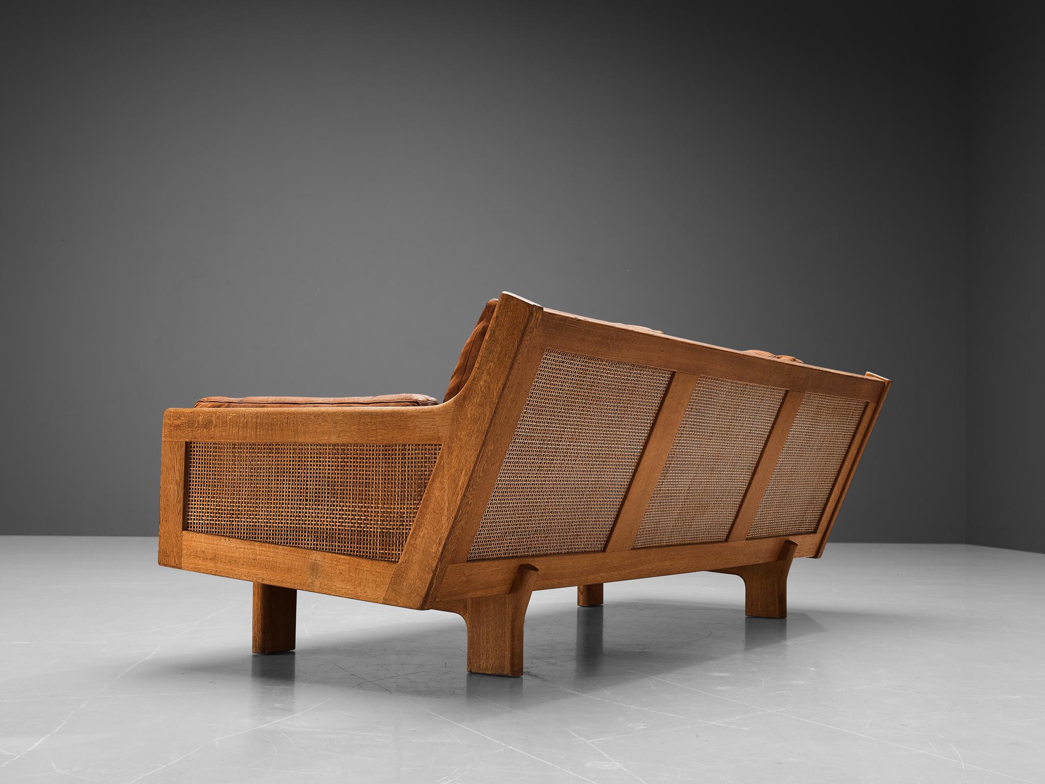 Three-seat sofa, oak, cane, leather, Denmark, 1960s 

This eccentric sofa features a splendid construction. When experienced from the side, the frame embodies a sharp angle that truly underlines the essence of the design. Both sides are executed in