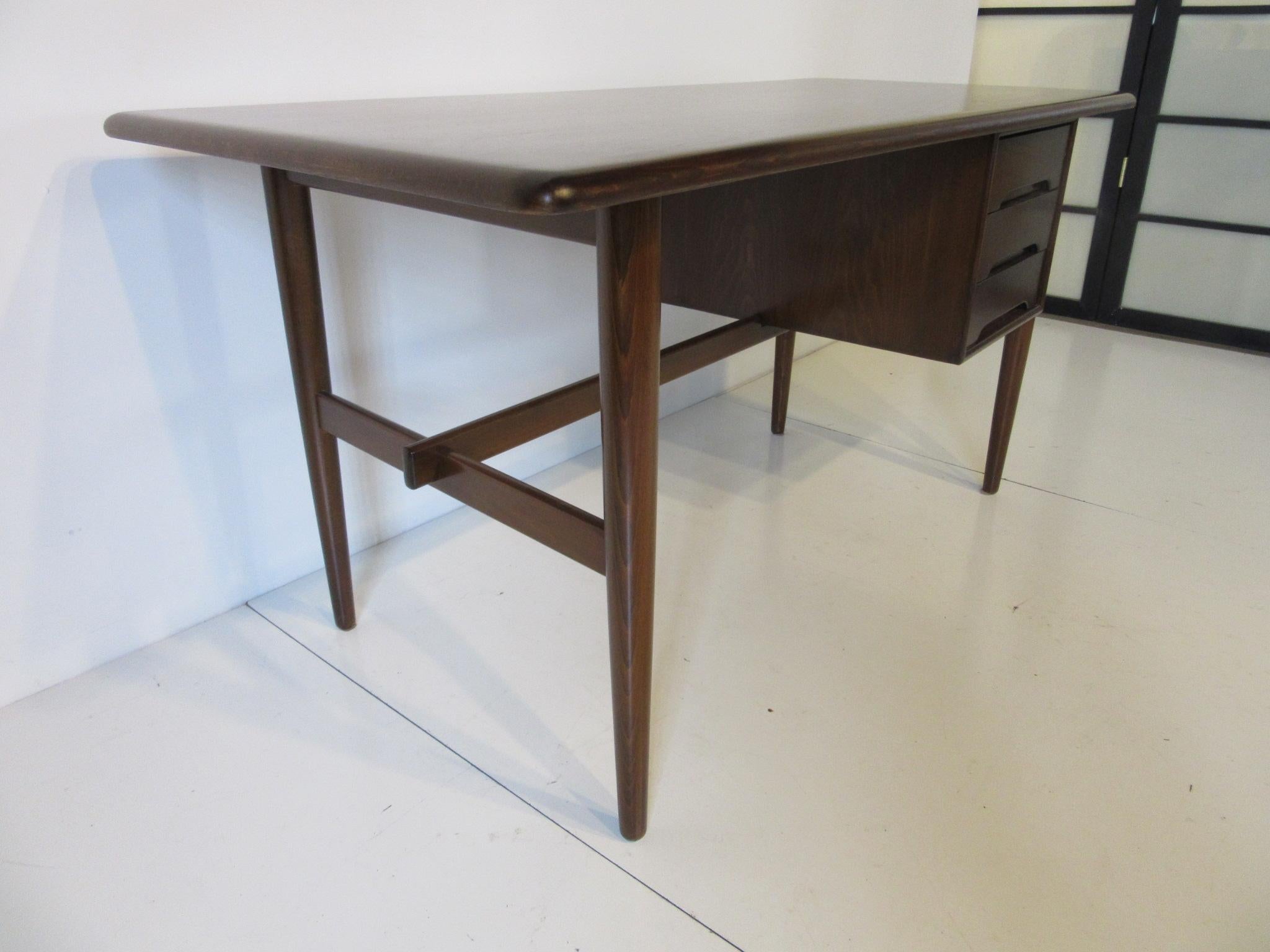 A dark Danish desk with angled top, three drawers to one side and cross stretcher to the lower portion for design detail and support. Branded mark to the bottom of the desk Made in Denmark, measurement for the angled desk top is 25