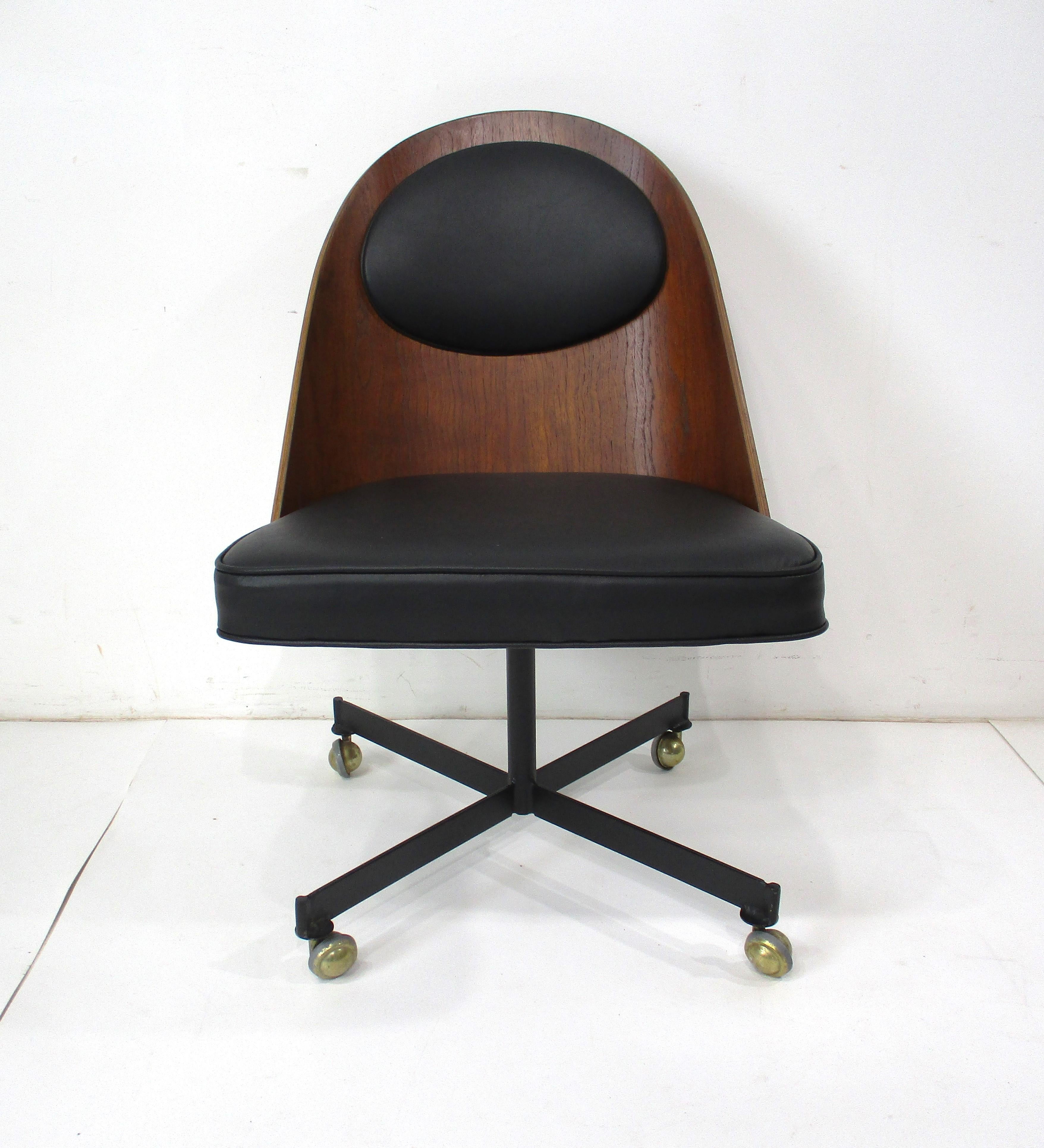 A very well crafted rolling desk chair with curved bent walnut back having top and bottom cushions in a soft black leather . The X base is in satin black steel with brass roller ball feet and the hardware to the back edge of the chair back has