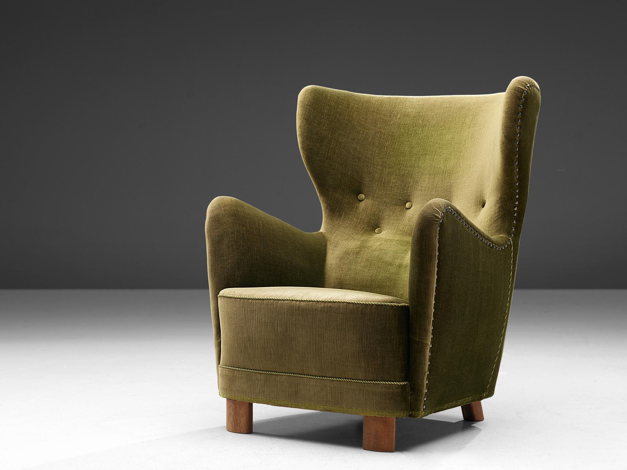 Wingback armchair in green upholstery, Denmark, 1950s.

The wingback chair is upholstered in a green velvet fabric and is finished with a green rope covering the straight seams. On the side of the chair the fabric is secured using furniture nails,