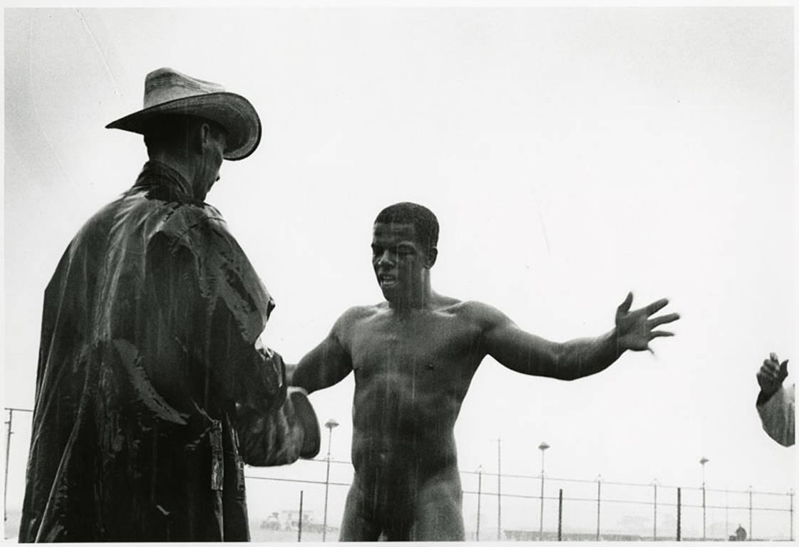 Shakedown, Ramsey Unit, Texas 1967-69 von Danny Lyon ist ein Schwarz-Weiß-Gelatinesilberdruck, der einen nackten schwarzen Mann zeigt, der vor einem Texas Ranger steht. Der Mann steht mit ausgebreiteten Armen und entblößt sich vor dem Polizisten.