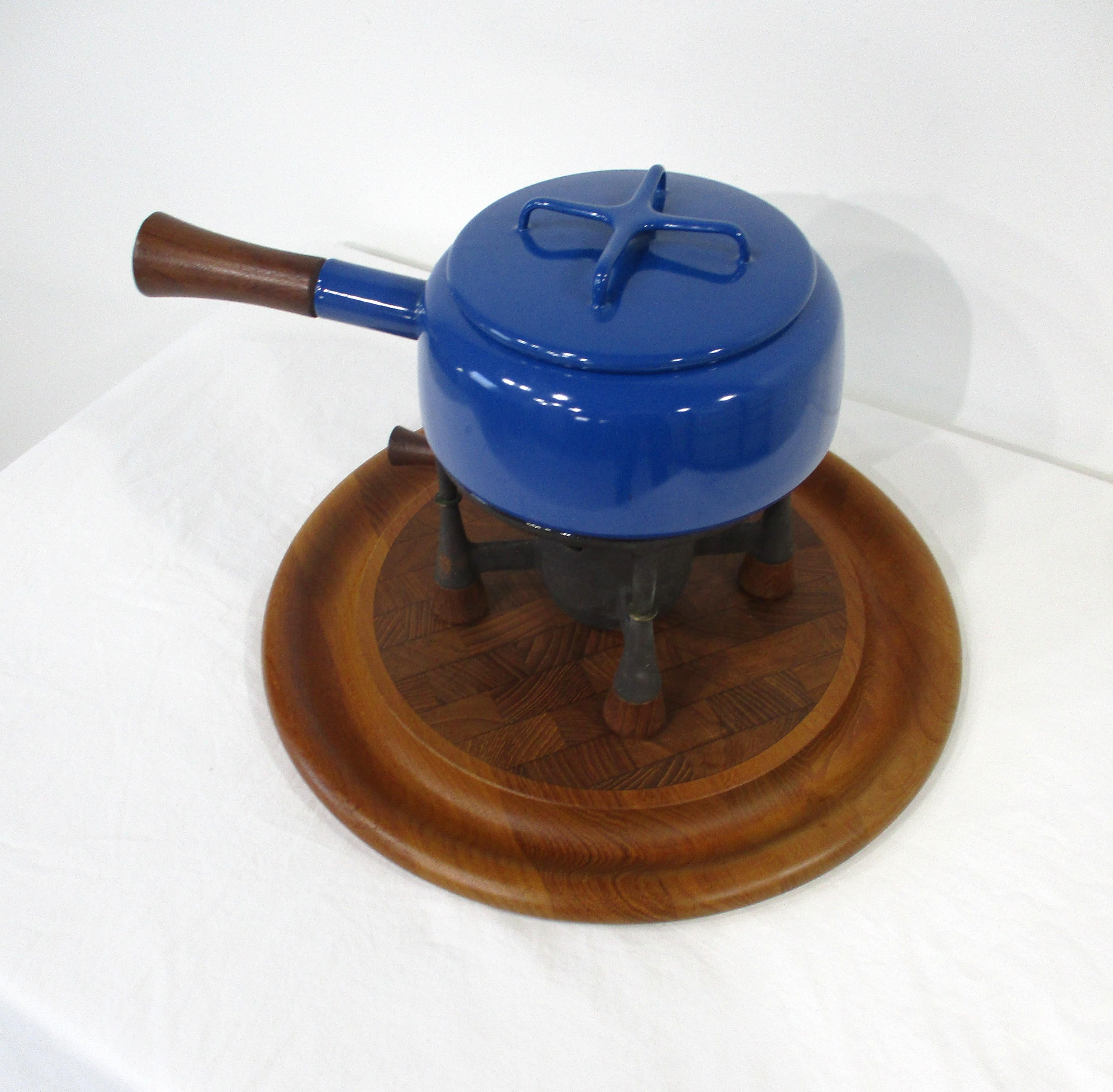 Un conjunto de tres piezas de utensilios de cocina o piezas de fondue en un azul brillante impresionante con olla y tapa , hierro y soporte de teca todo sentado en una bandeja de teca . Fabricado por Dansk conocido por sus materiales de alta calidad