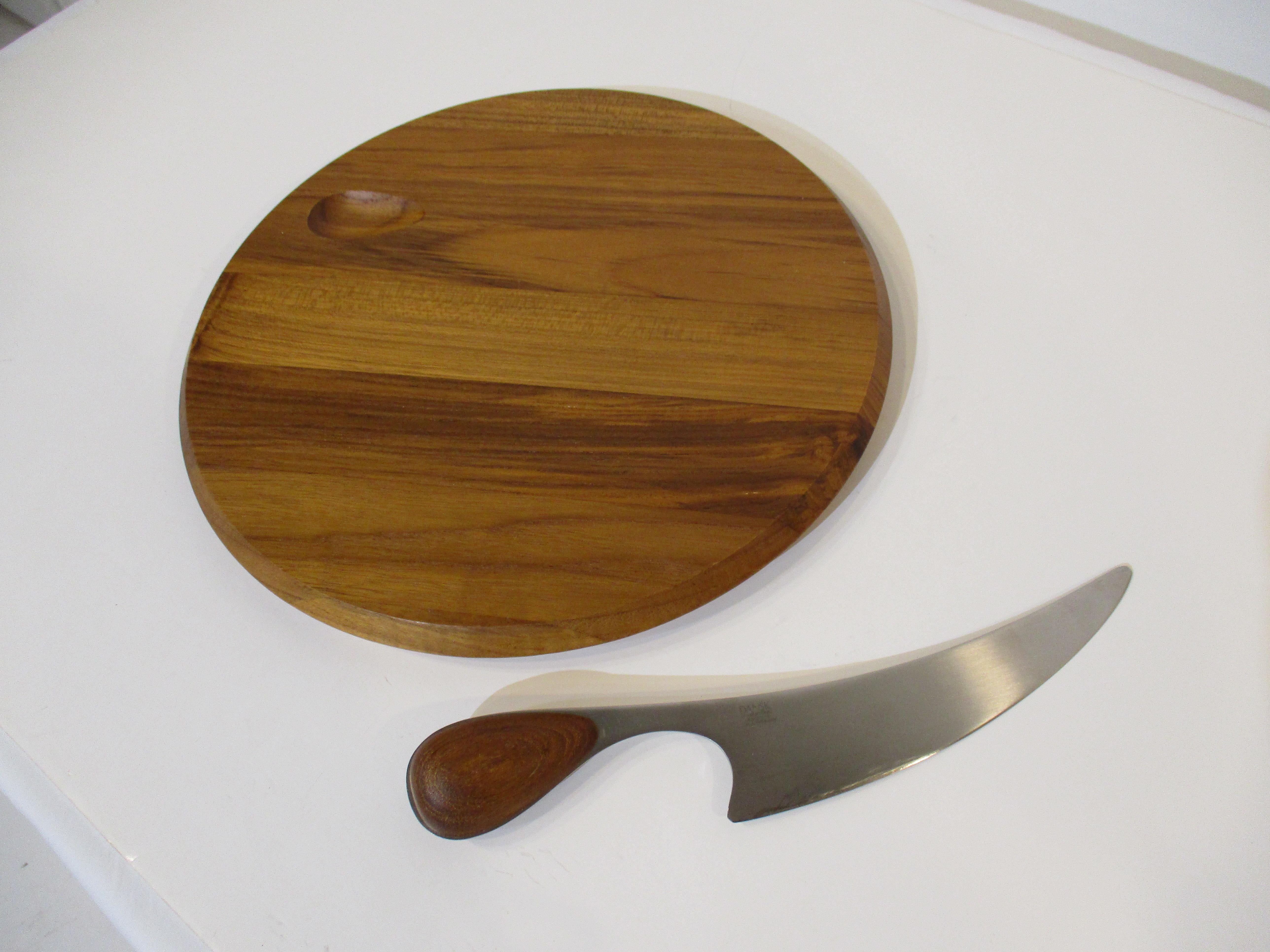 A very well crafted staved teak wood cheese board with carved out area to the top edge of the board for the handle of the knife to sit . The large stainless steel curved bladed knife has a matching teak handle and the boards edge is tapered and