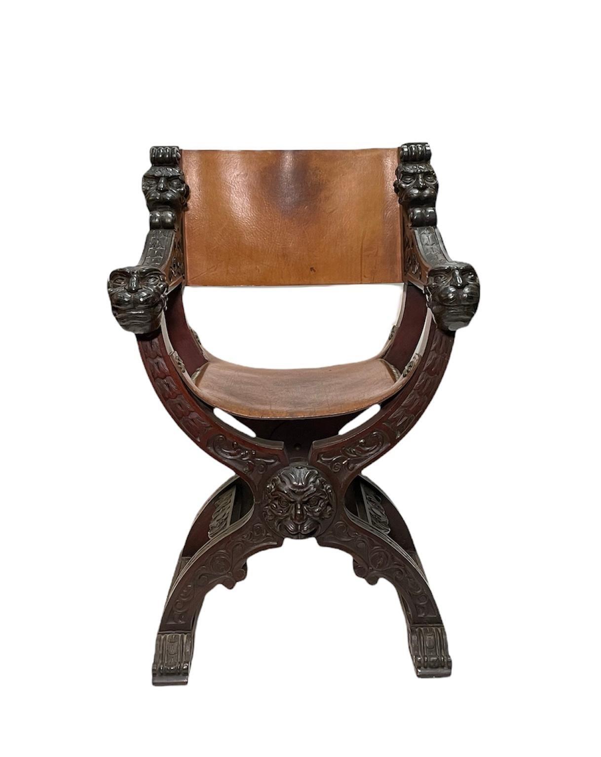 This is a Dante or Savonarola wood and leather heavy chair. This is an X- shaped frame chair. The whole chair is decorated with a very detailed carving of a mix of a lion-man face head masks in the rest arms, back and in the frontal center of the