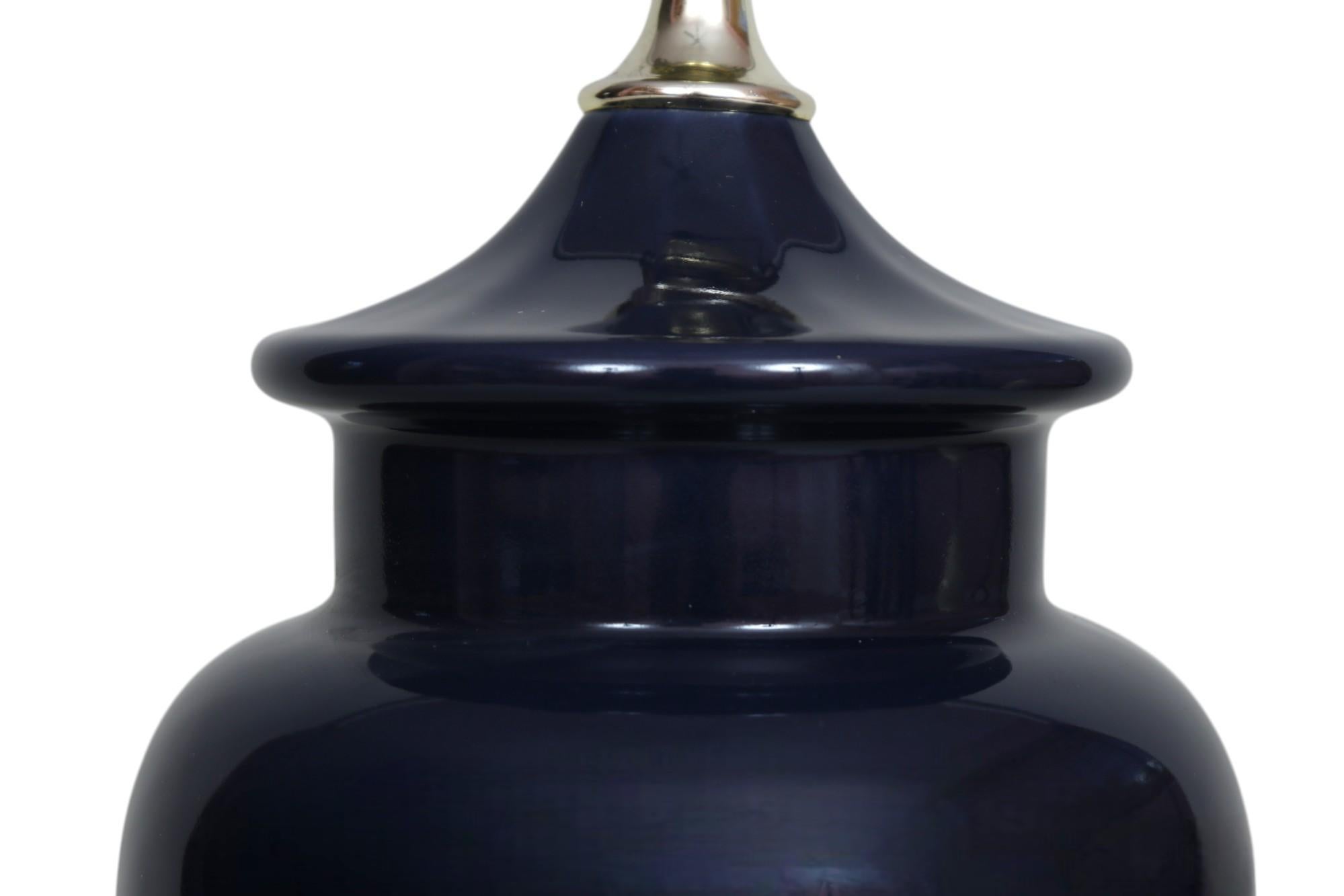 A deep blue ceramic table lamp. The round ceramic vase is ginger jar shaped, and sits on a round beveled wooden base with ming feet. Measures 21.5