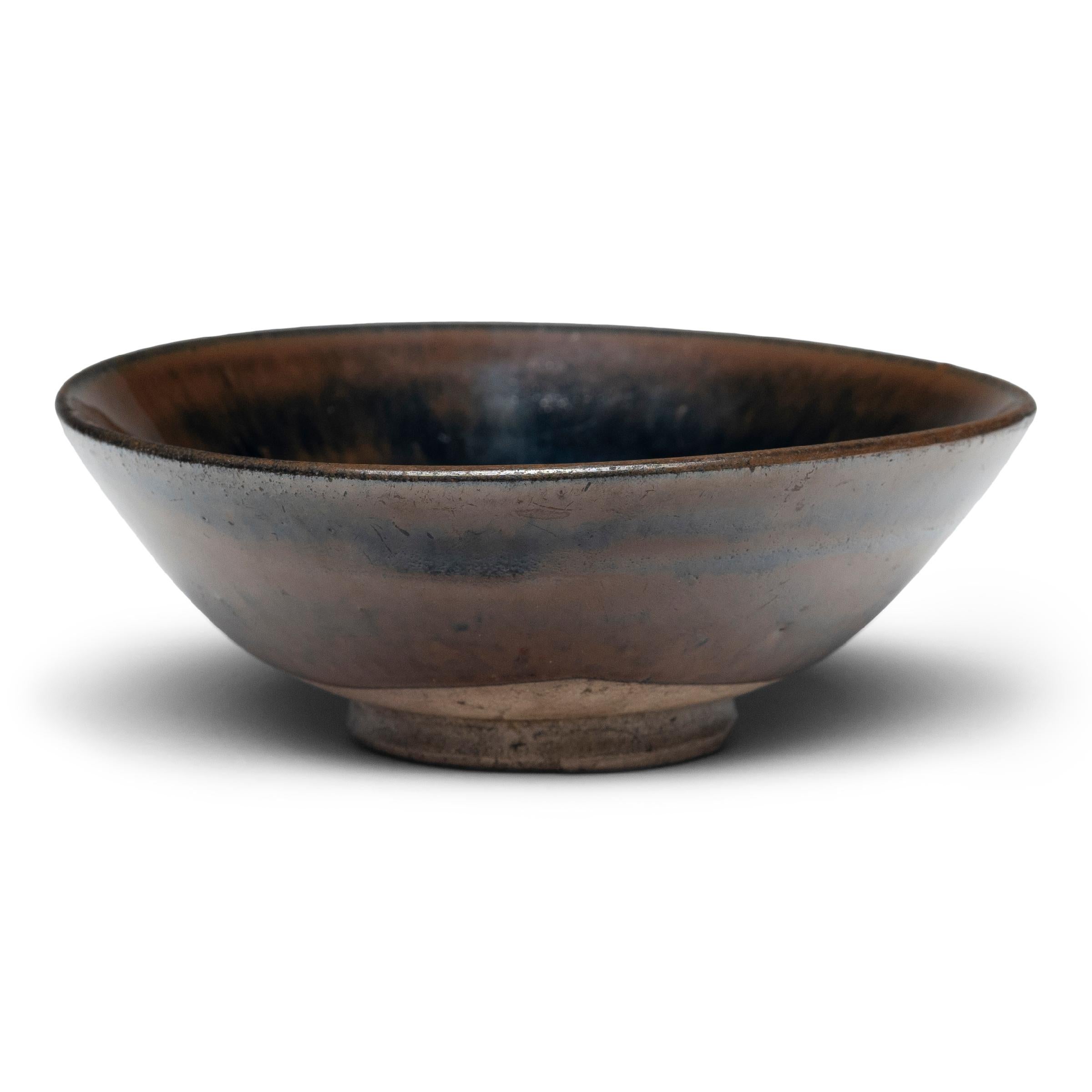 This darkly glazed stoneware bowl is simply shaped with tapered sides and a footed base. A rich, glossy glaze coats the bowl inside and out, streaking down the sides to reveal a depth of color ranging from light brown to dark green to inky black.