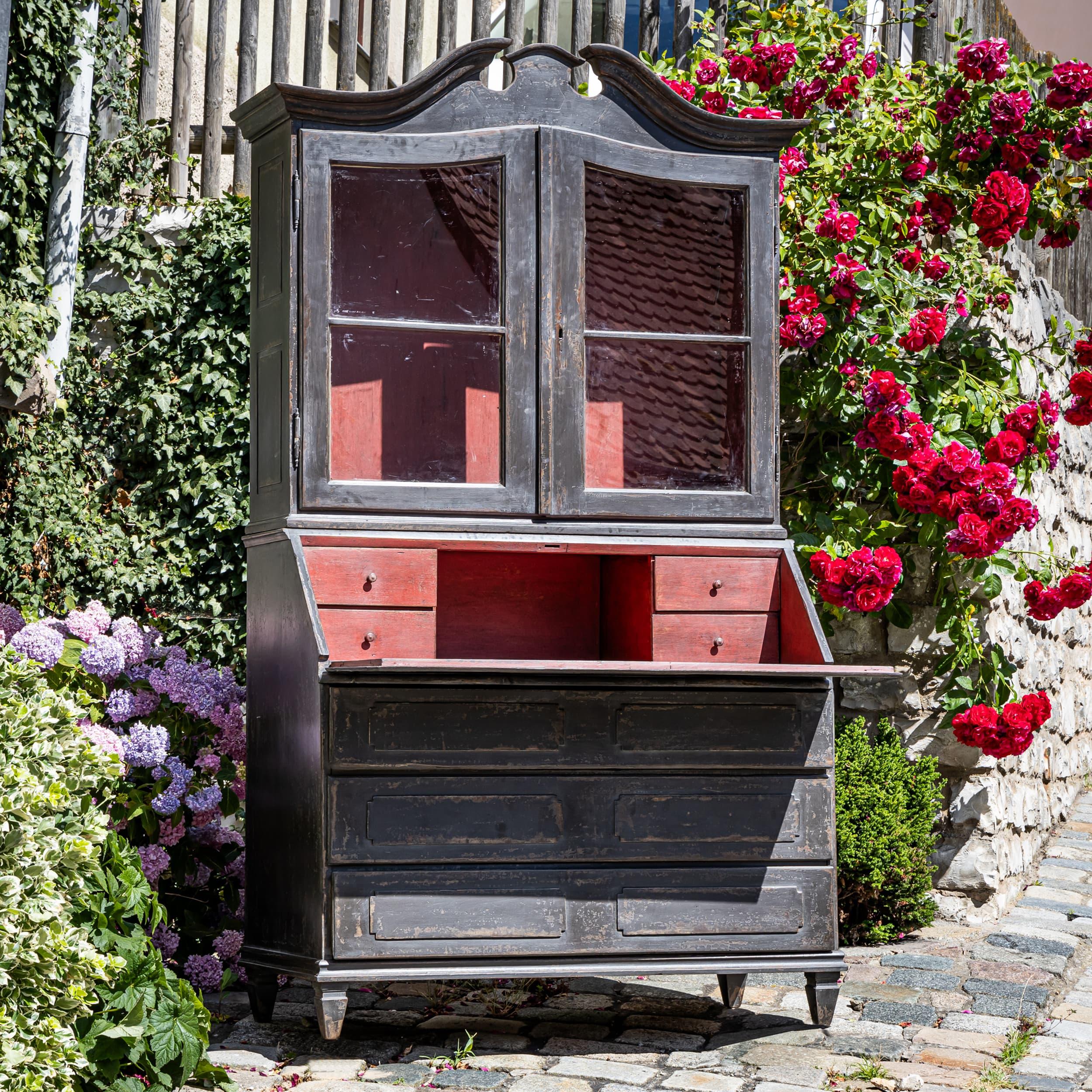 Baroque secretary cabinet featuring a two-door glazed display hutch above and a lower section with a slant-front writing desk and three drawers. The dark gray paint finish is newly applied, with a contrasting interior in brick red for a harmonious