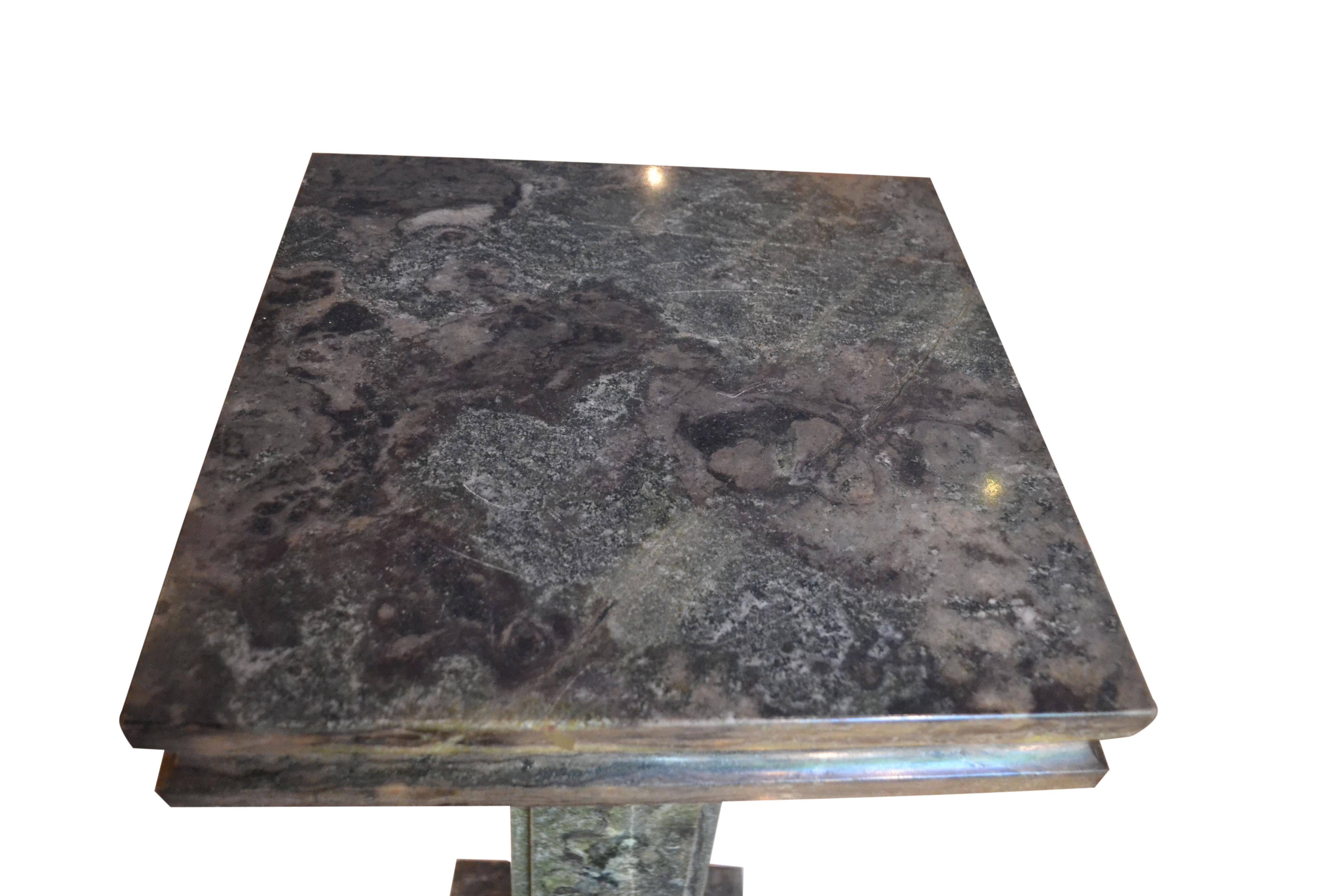 A tall dark green/grey marble column; the rectangular column supports a larger tiered square marble top; the bottom of the column sits on a patinated bronze decorative base on a further shaped rectangular base. 
 