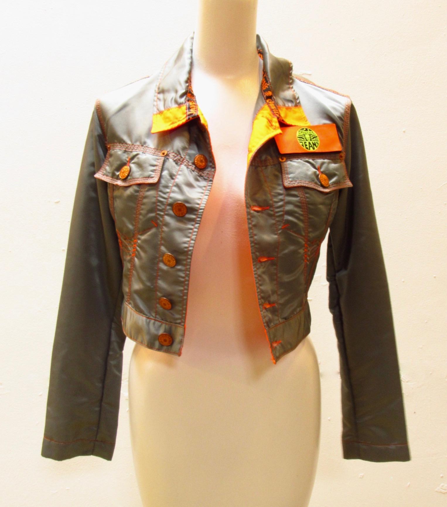 Cropped dark grey nylon jacket from vintage Jean Paul Gaultier is a standout with its orange seams, contrast stitching, and removable velcro JPG patch. Sleeves feature columns of buttons and back boasts a buckle that allows for an adjustable fit.  