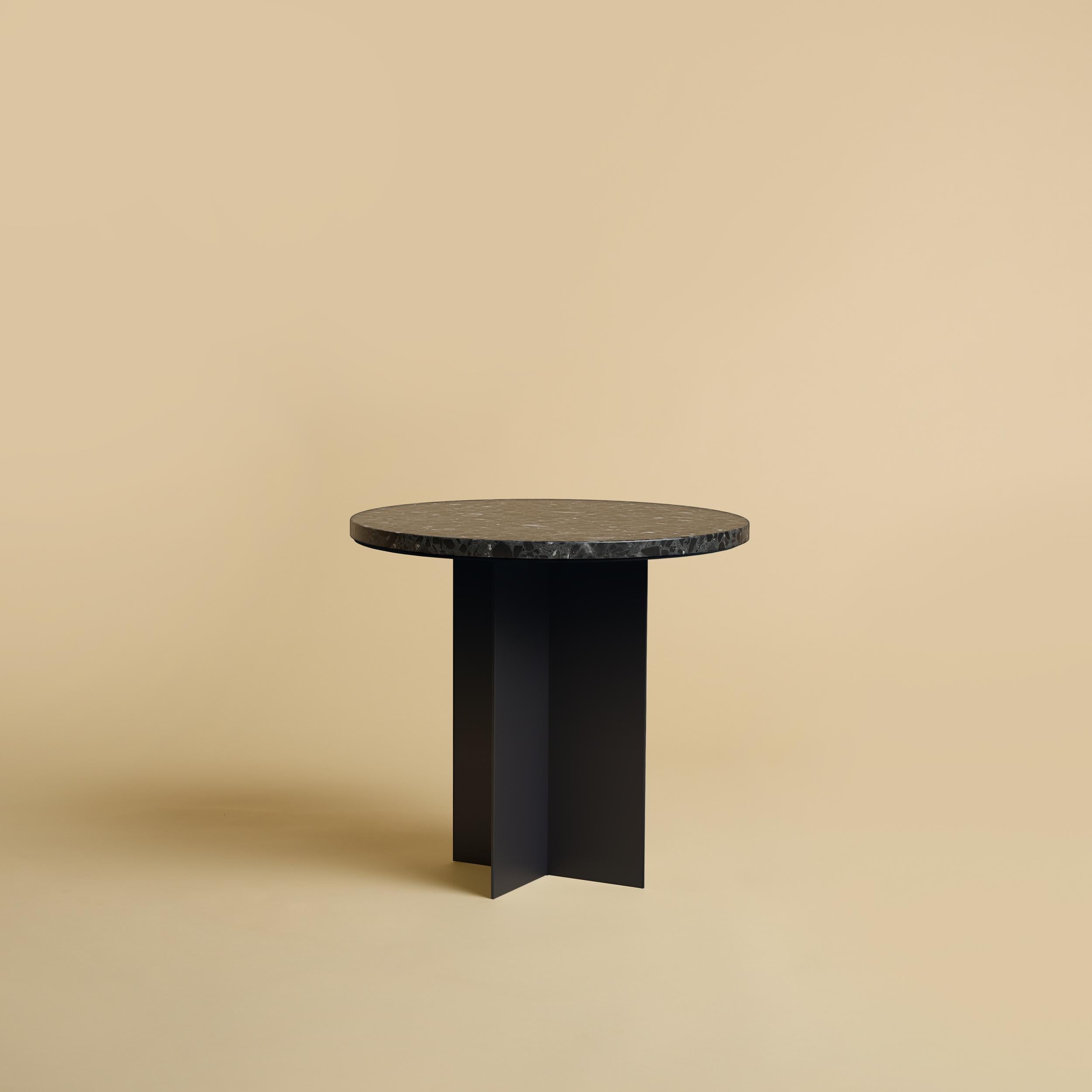La table basse Baker est fabriquée avec une base en métal revêtu de poudre et un plateau en terrazzo Mystery gris foncé. La partie supérieure, de couleur gris foncé, est circulaire et a un diamètre de 45 cm, tandis que la base est obtenue en collant