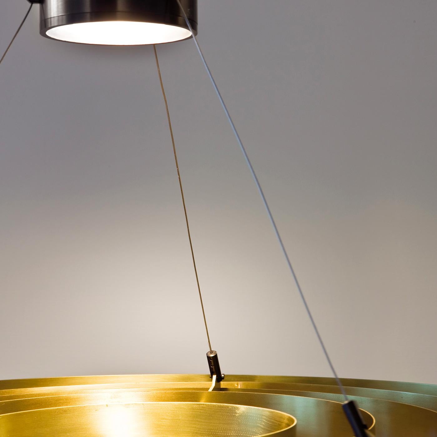 This elegant lamp consists of a central lighting body suspended from the ceiling thanks to steel cables. Below it, a series of concentric brass rings create a striking optical effect.
  