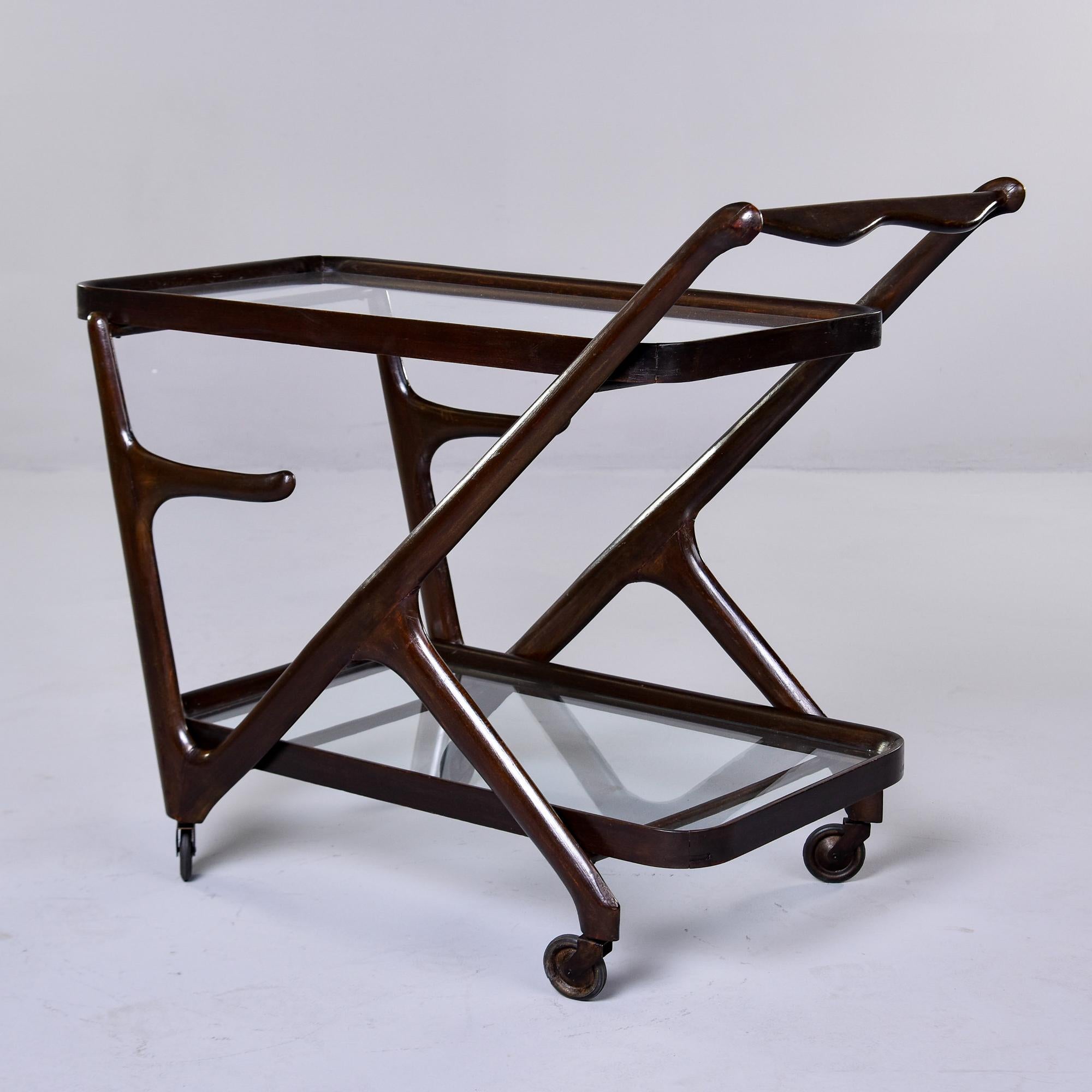This modernist style bar cart was designed by Cesare Lacca for Cassina. It has a dark mahogany frame, two glass shelves and dates from the 1950s. Original metal and black rubber casters, angled frame and two tiers of glass insert shelves. 

Overall