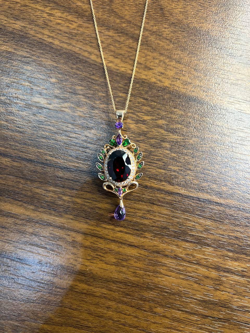A striking pendant necklace featuring a central red garnet. Set in a delicate gold frame, this piece is adorned with an array of colorful tsavorites, diamonds and others encircling the central gem, creating a vibrant halo effect. The pendant is