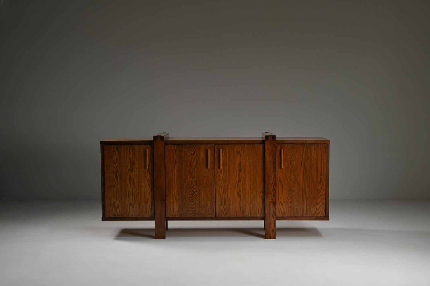 This Belgian sideboard is crafted mostly from solid oak and reflects the Brutalist style with its strong lines and architectural details. The piece offers plenty of storage with four cabinets. Its dark stained oak finish brings warmth and simplicity
