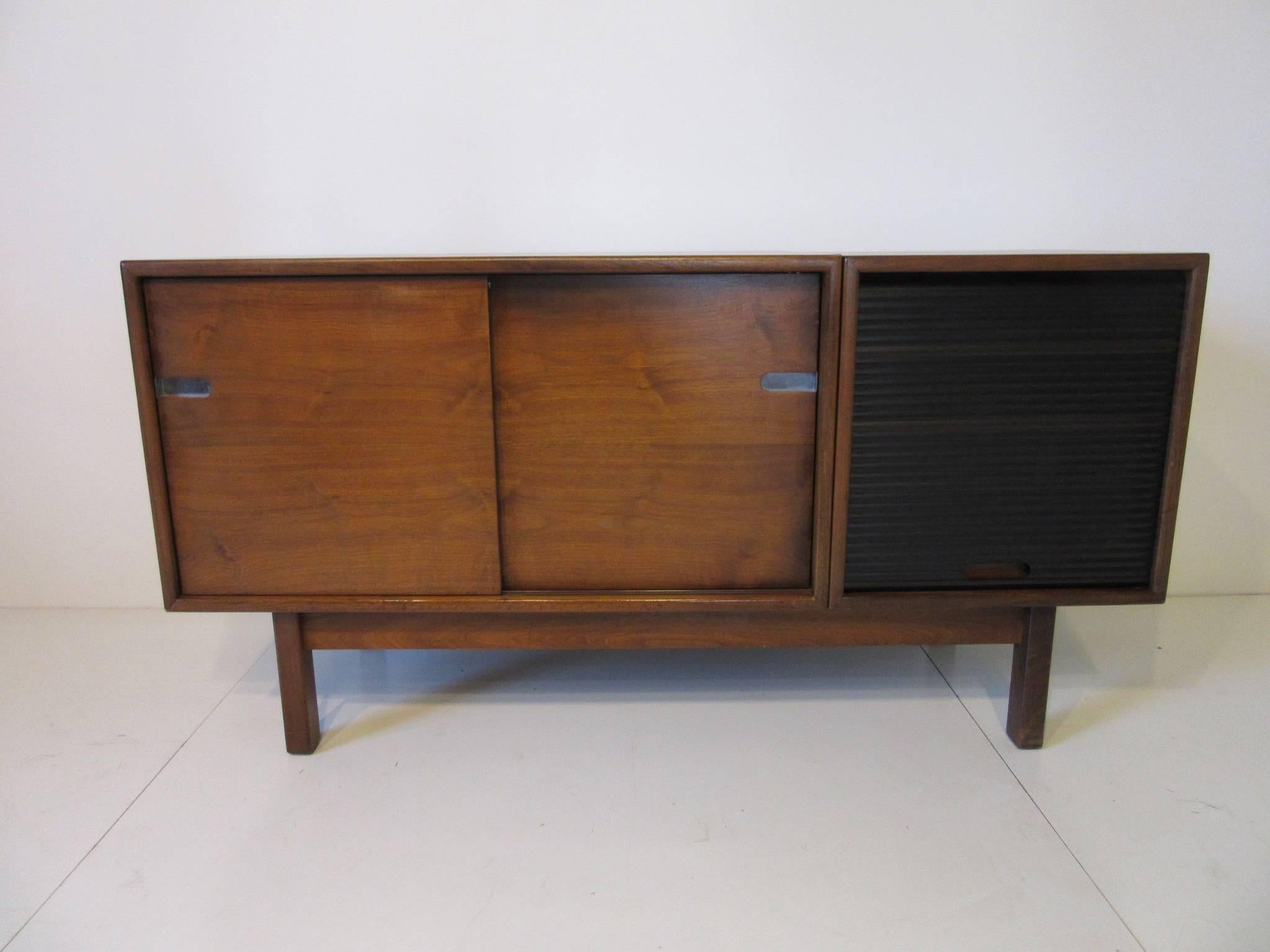 A very well crafted dark grained walnut credenza with double sliding doors and a dark anodized metal pull up tambour door with three drawers. Inside the sliding doors Revel two adjustable shelves and detailed cutout door pulls inset with polished