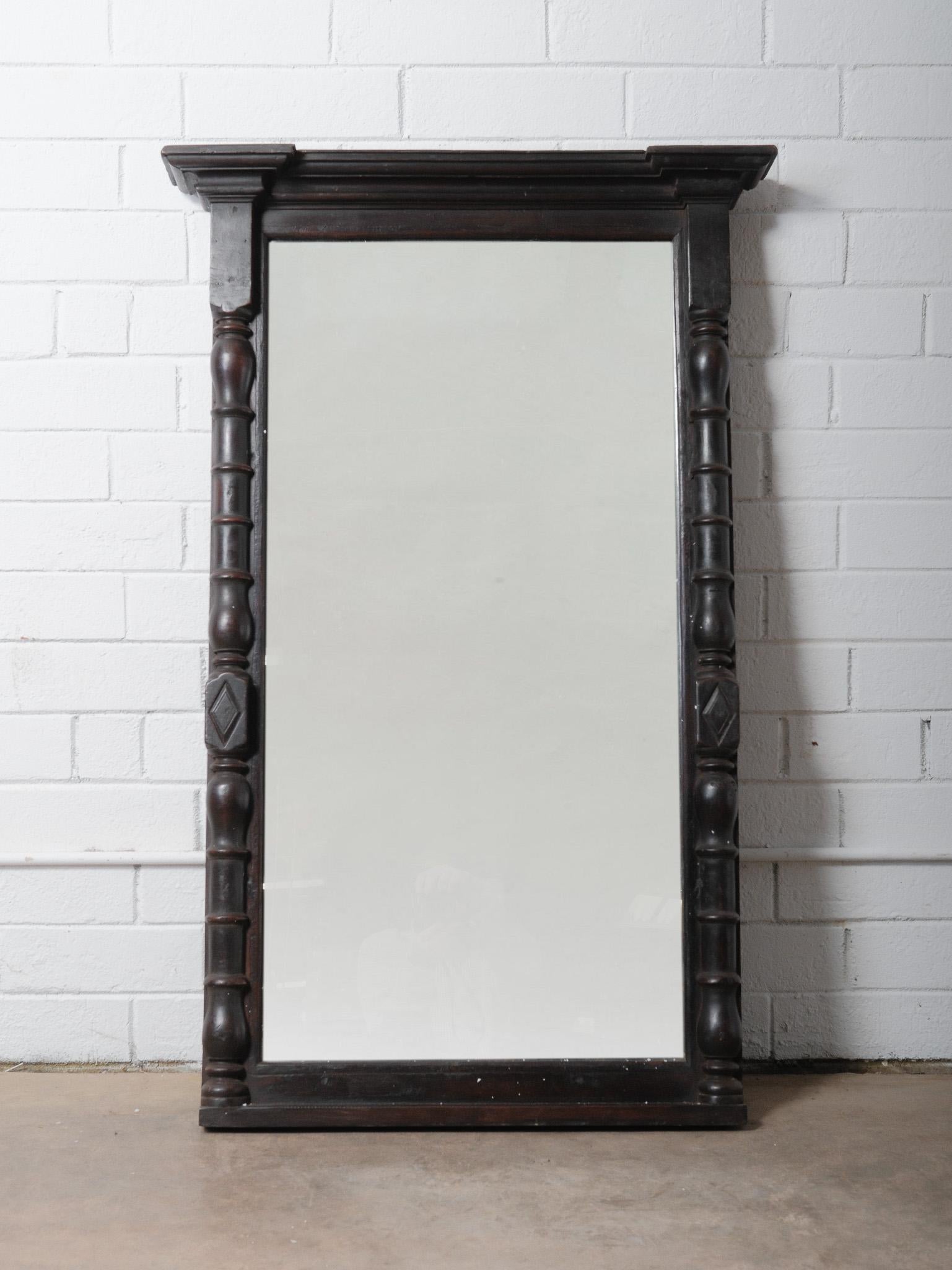 This wooden mirror frame from India showcases a beautifully aged dark finish and 20th-century craftsmanship. The frame features hand-turned column detailing along both sides, adding sculptural depth and a sense of classical elegance. Its crown