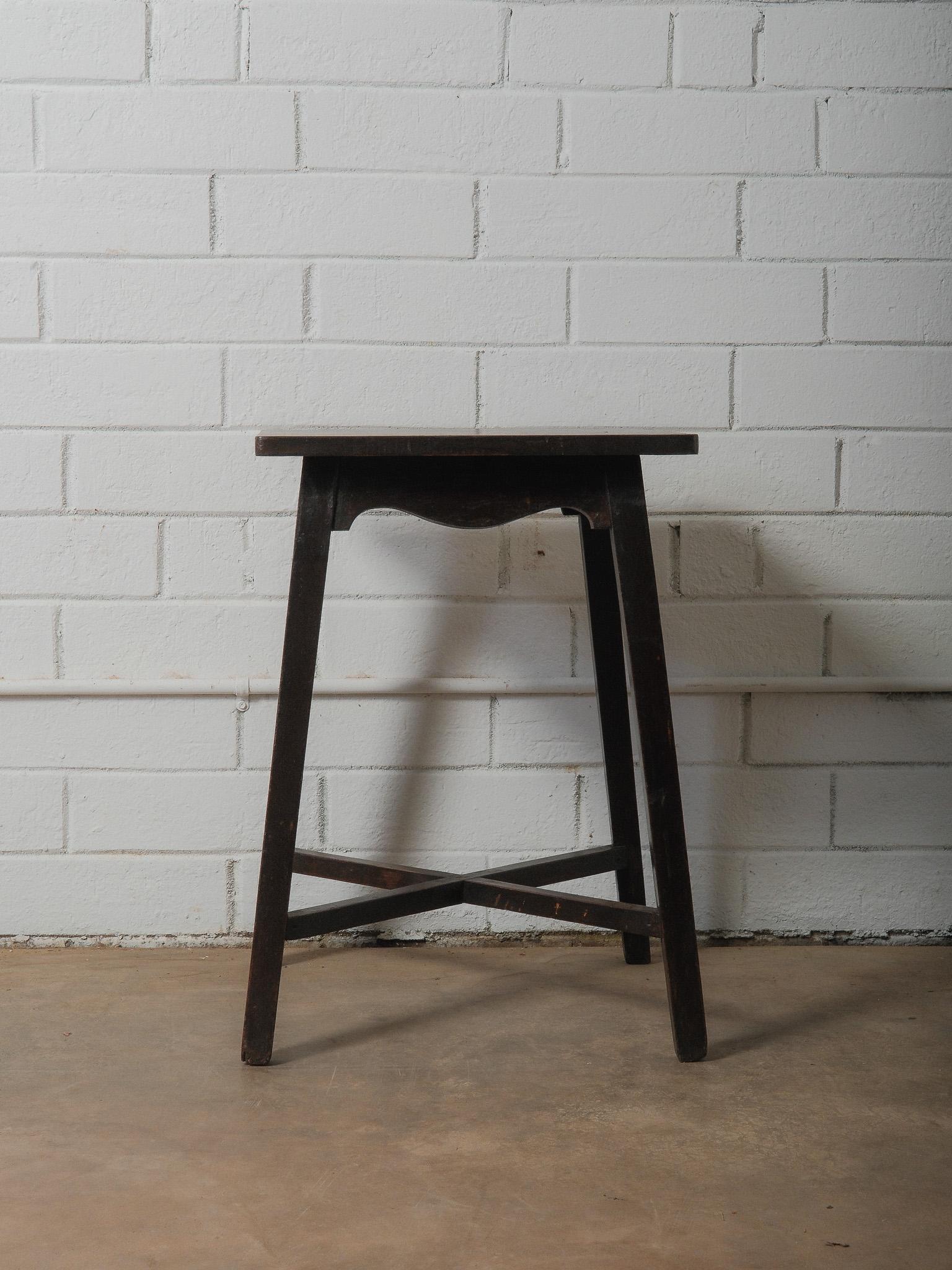 A wooden table with a darkened surface and slender proportions. The rectangular top rests on four splayed legs, joined by low stretchers that cross at the center for stability. The apron is shaped with a subtle curved profile, adding a quiet