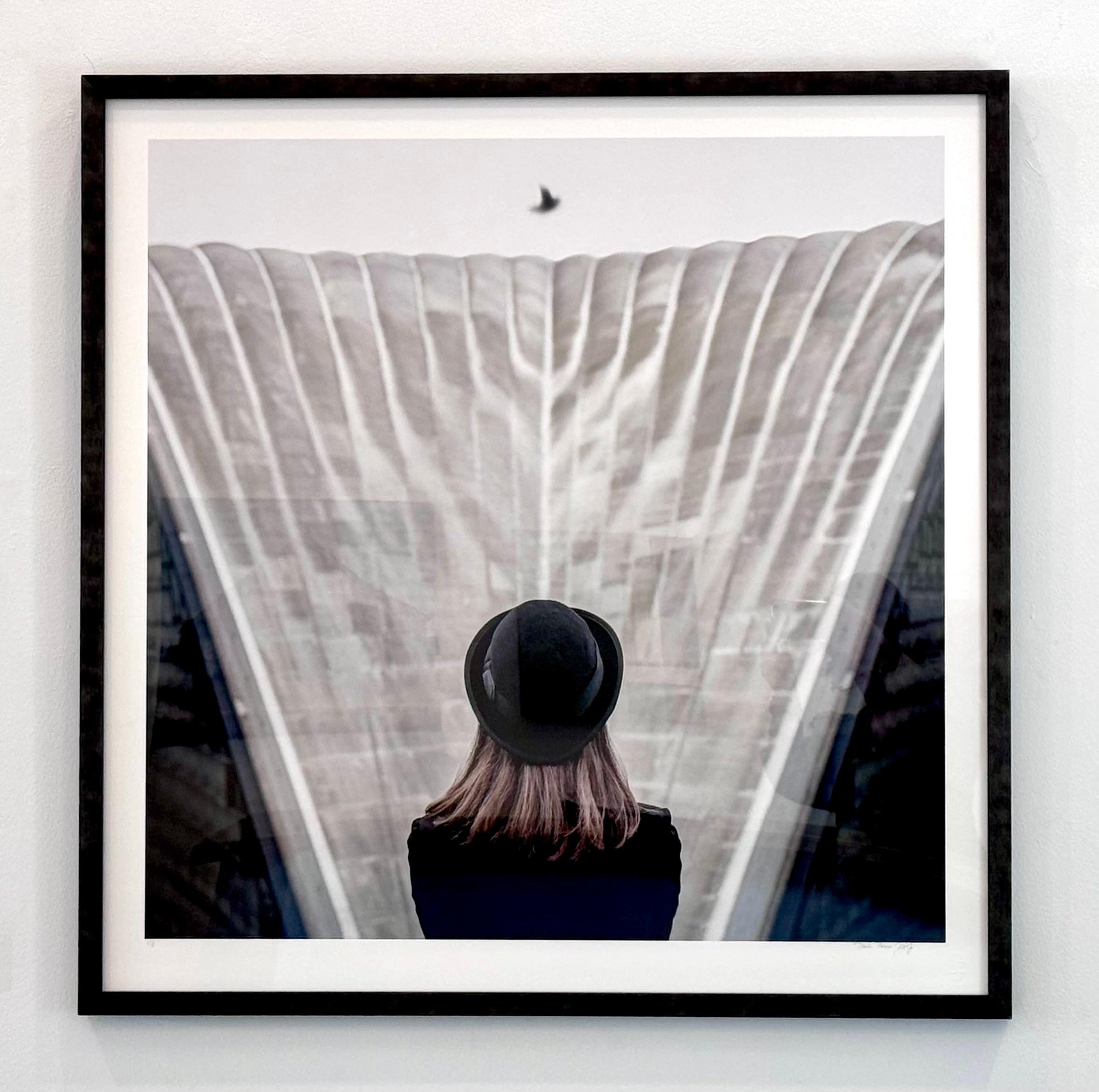 Photographie couleur encadrée d'une femme avec un chapeau noir de style Magritte, architecture grise - Noir Color Photograph par Dasha Pears