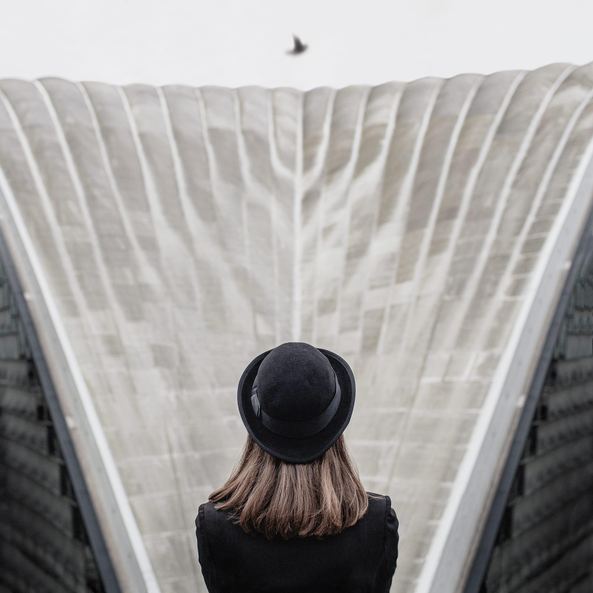 Gerahmte Farbfotografie einer Frau mit schwarzem Hut im Magritte-Stil, graue Architektur