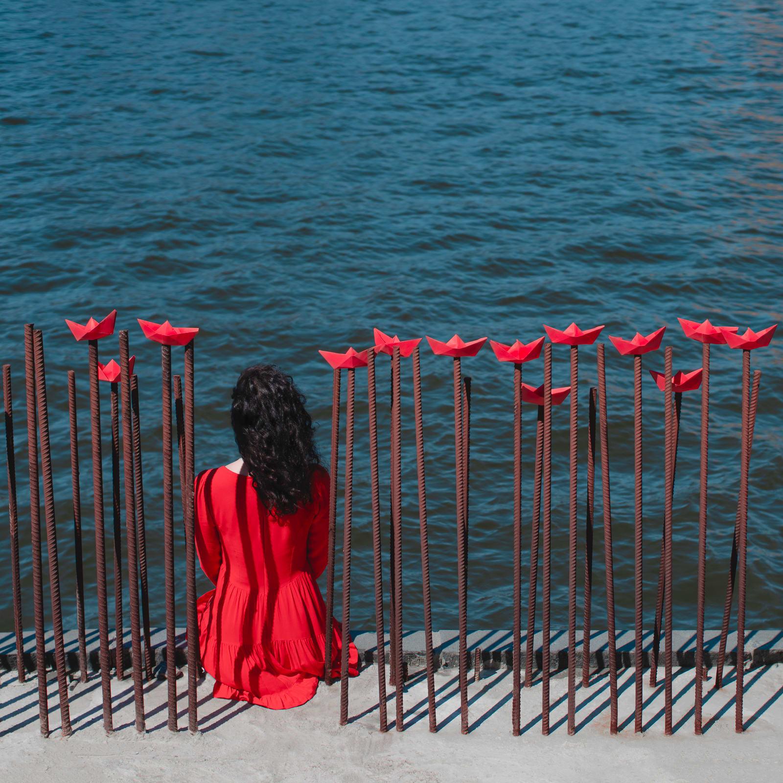 This beautifully vibrant landscape features the back of a woman in red, gazing out at the blue ocean. Little red origami boats adorn posts around her. Will she sail away? This is a limited edition color photograph. Number 1 of 5 is currently
