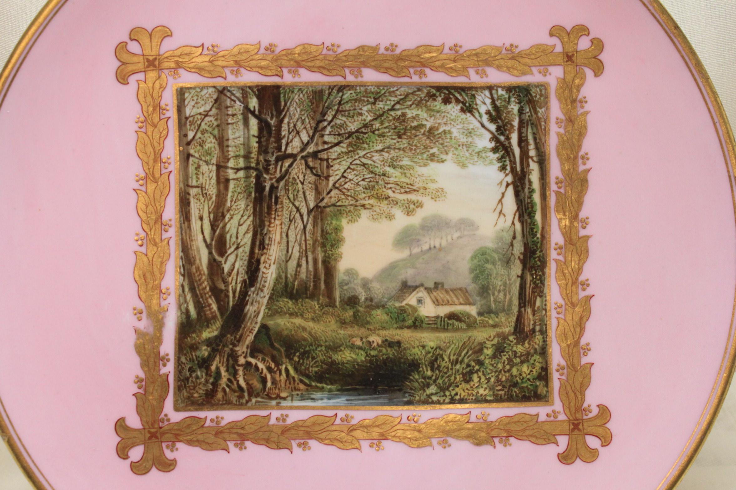 The centre of this Davenport porcelain plate is decorated with a hand painted scene of a farmhouse on the edge of a dense wood, with sheep grazing in a meadow in the foreground, on the edge of a stream. Surrounding this idyll is a gilt frame in the