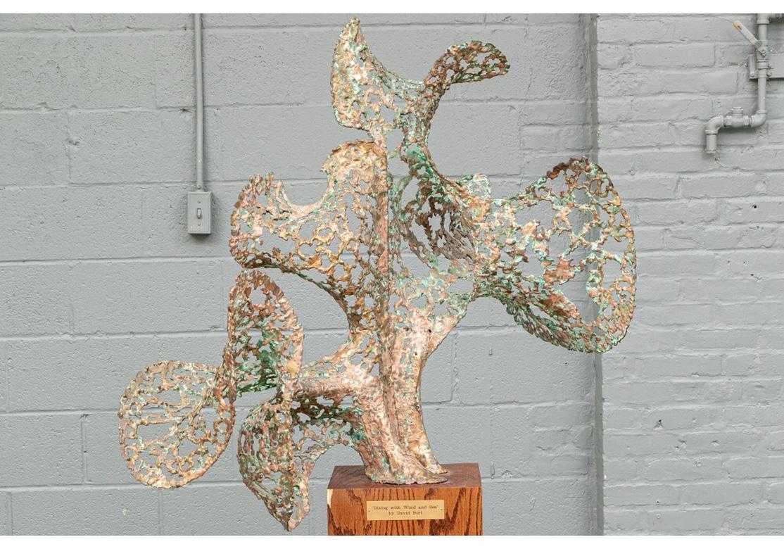 Targa sulla base del piedistallo in legno impiallacciato con artista e titolo. Una scultura astratta in rame traforato di grandi dimensioni con un ampio disegno cuvilineo a vento, dalla forma quasi simile a quella di un uccello. L'artista è stata