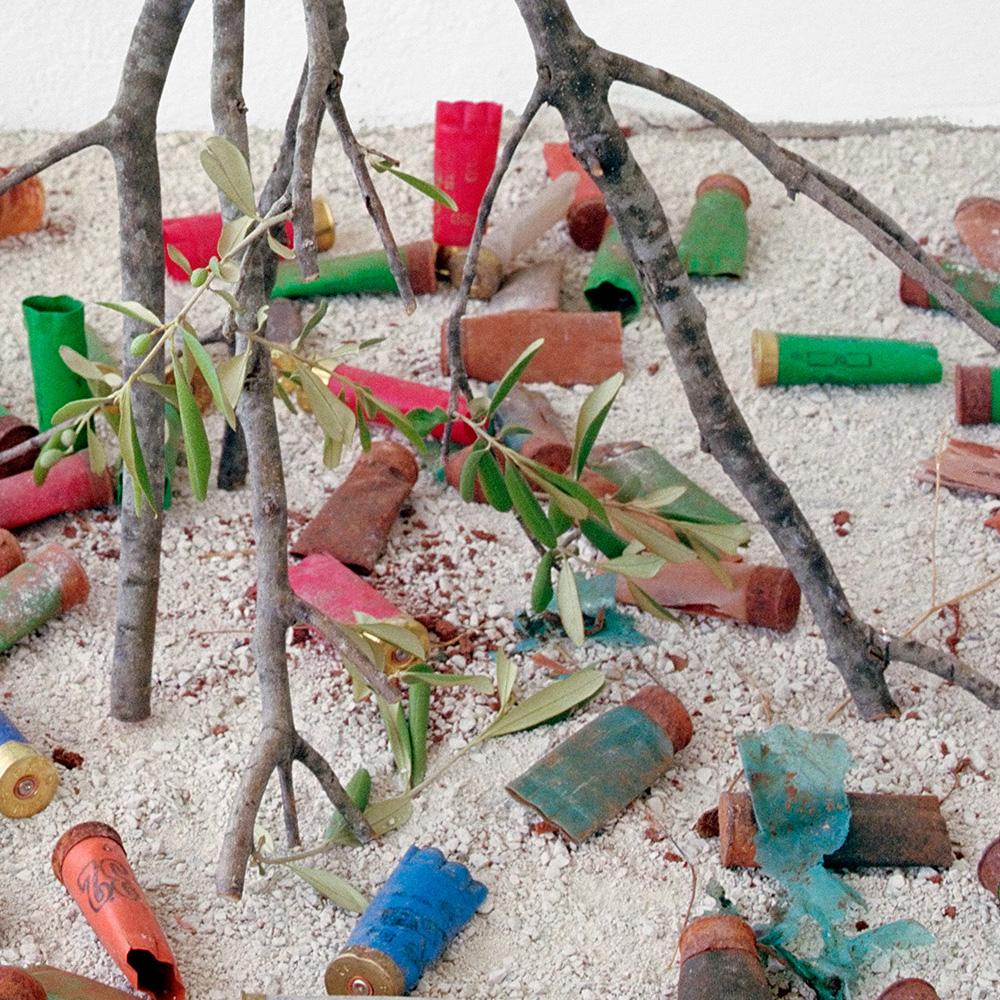 Color still life photograph of a figurative olive branch, brick, chestnuts, and shotgun shells
