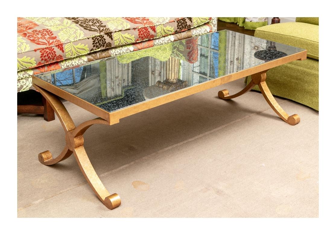 A great looking cocktail table in the manner of the Hollywood Regency style. The metal frame in his antique gold finish with curved X shaped supports on scrolled feet. With an inset mottled glass top. 
Condition: There are some deep scratches to the