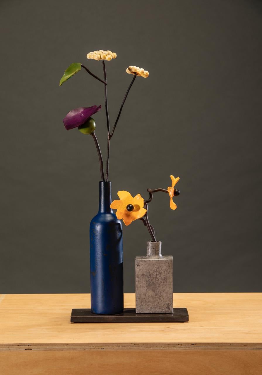 'Blue Bottle Summer' by David Kimball Anderson, 2021. Bronze, steel, and paint, 21 x 12 x 10 in. This sculpture features two vases cast in bronze and finished in patinas of blue and grey, respectively. In the blue vase, one flower is painted in