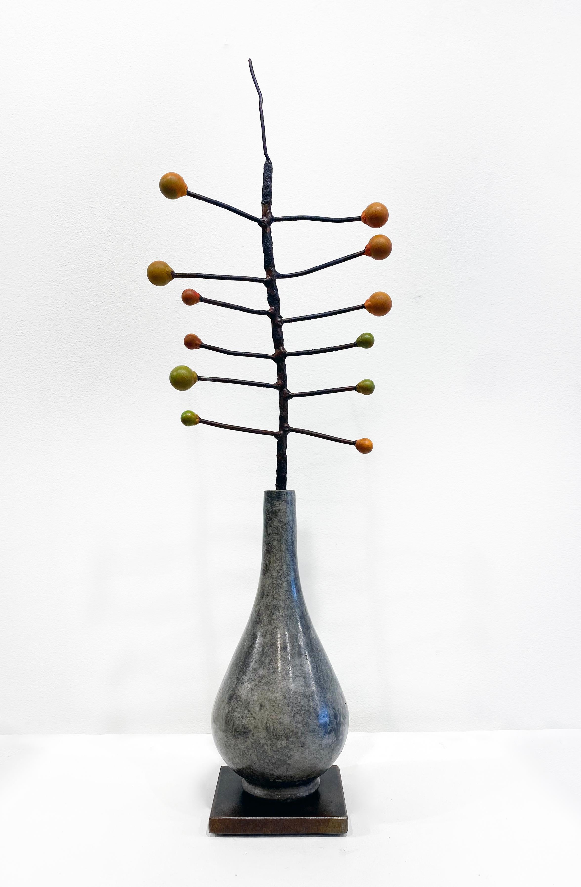 'Seeds' by David Kimball Anderson, 2022. Bronze, steel, and paint, 24 x 8 x 6 in. This sculpture features a classic rounded vase cast in bronze and finished with a grey patina. It features a series of bronze branches with colorful green and orange