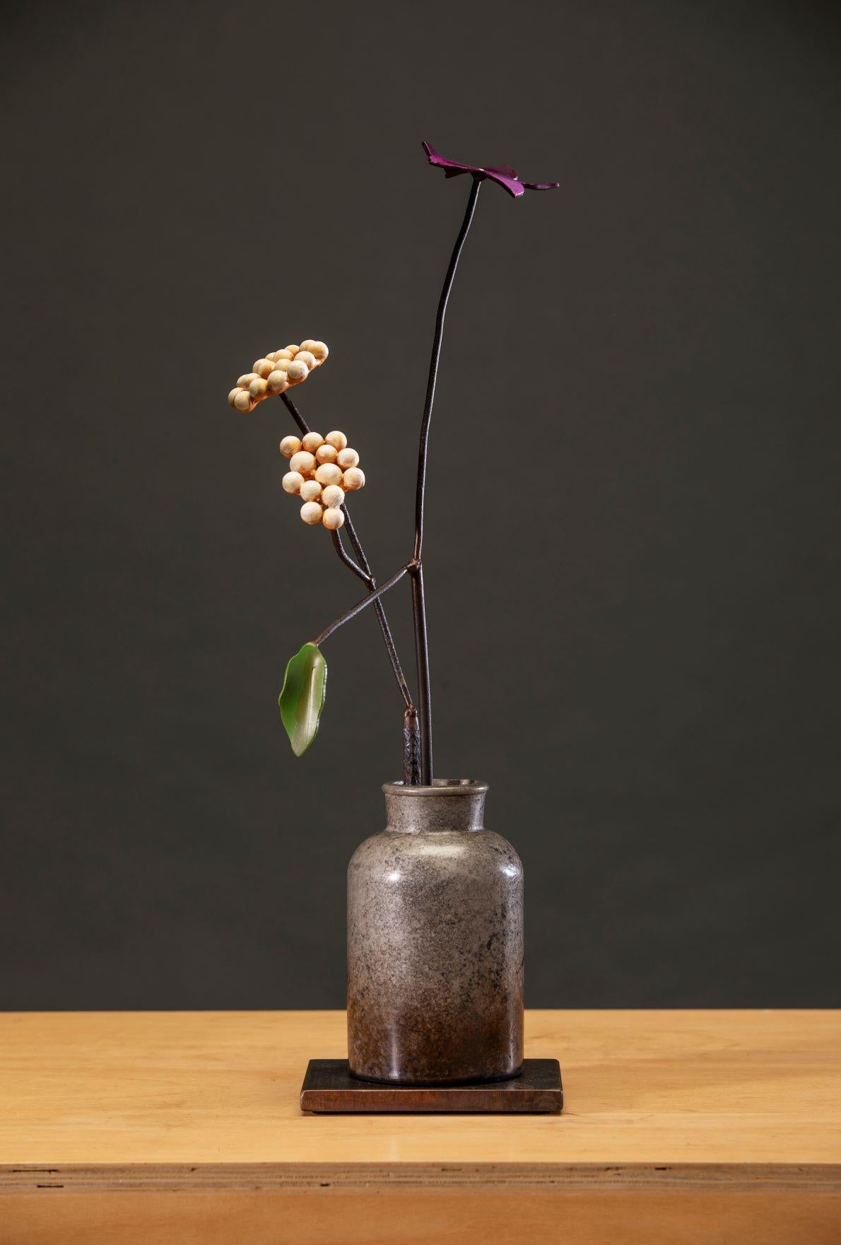 'Yarrow in a Dutch Jar' by David Kimball Anderson, 2022. Bronze, steel, and paint, 23 x 7 x 7 in. This sculpture features a round, cylinder vase cast in bronze and finished with a grey patina. It features a steel flower painted in purple with two