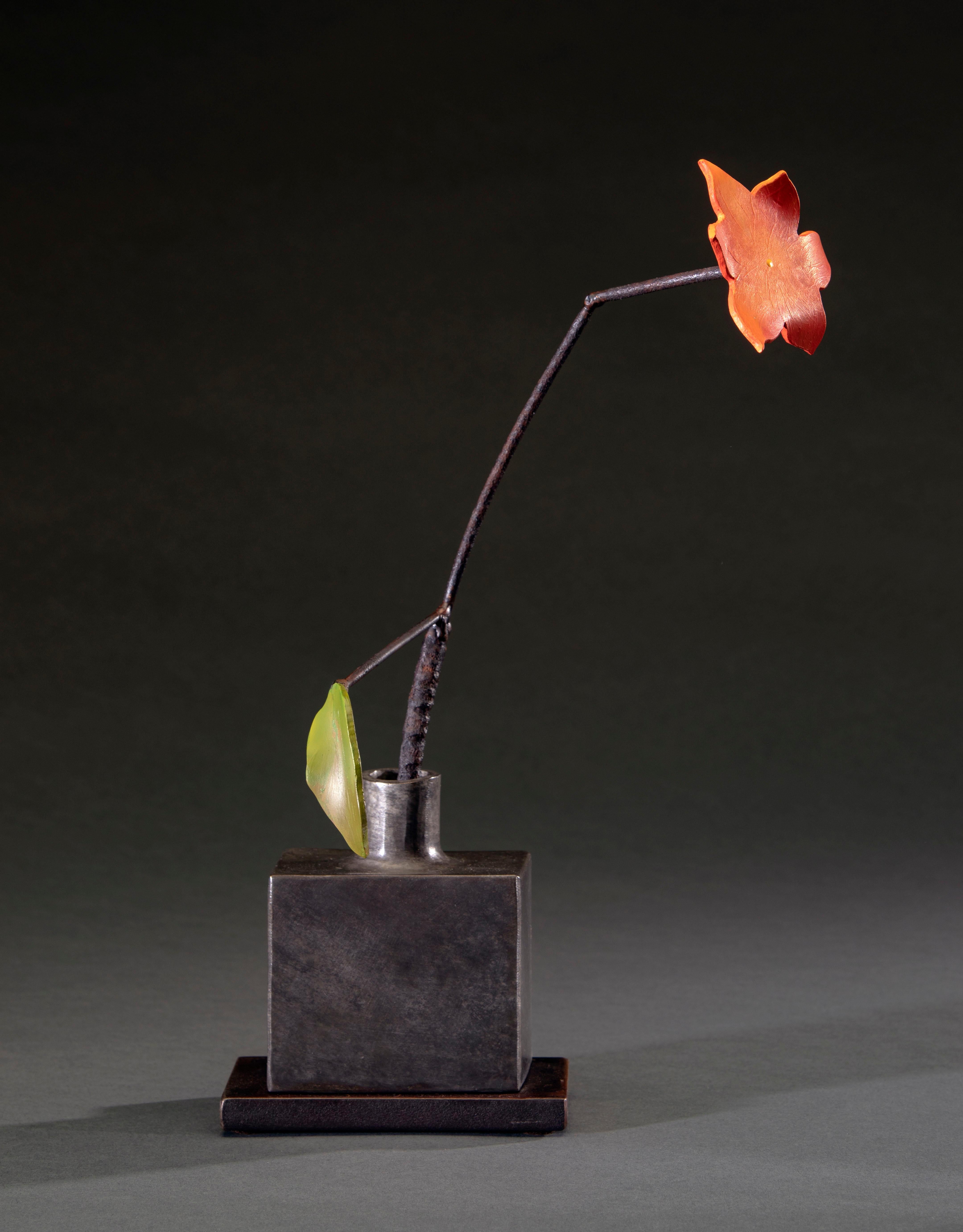'Silver Bottle Orange Flower' by David Kimball Anderson, 2022. Bronze, steel, and paint, 18 x 10 x 5 in. This sculpture features a  square vase cast in bronze and finished with a grey patina. It features a steel flower painted in bright orange and a