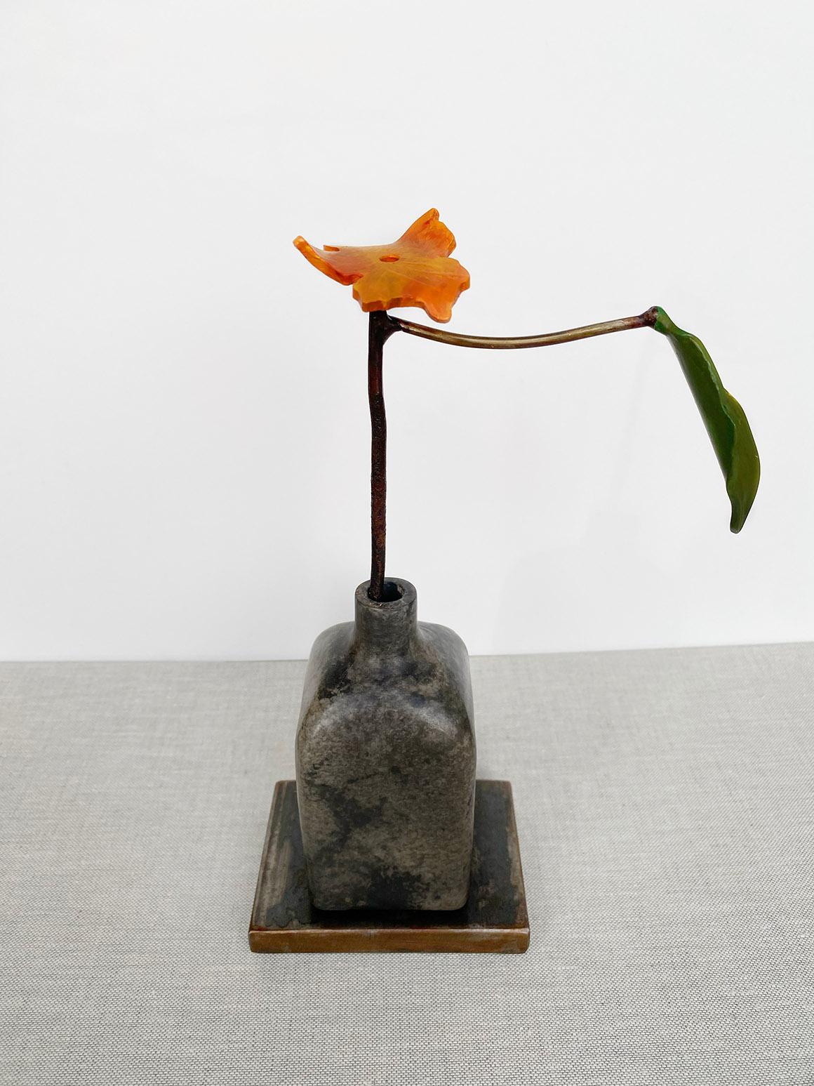 'Persimmon' by David Kimball Anderson, 2020. Bronze, steel, and paint, 10 x 4 x 3 in. This sculpture features a round vase cast in bronze and finished in varying patinas of light and dark gray. The flower is in a bright color of orange and 1 green