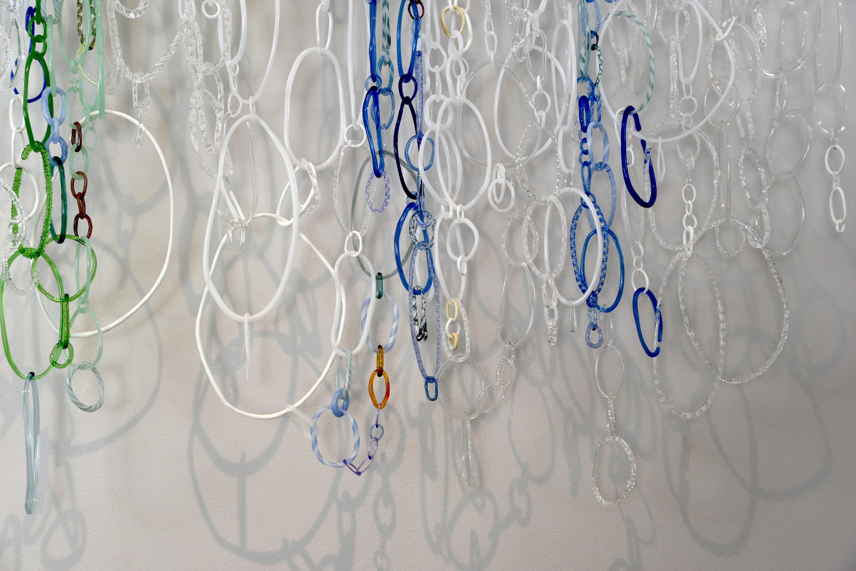 This hanging sculpture by David Licata is composed of free-form circular and oval loops of torch-worked borosilicate glass in white, clear, green, cobalt and periwinkle blue. The loops, in varying sizes and textures, cascade downward from a white