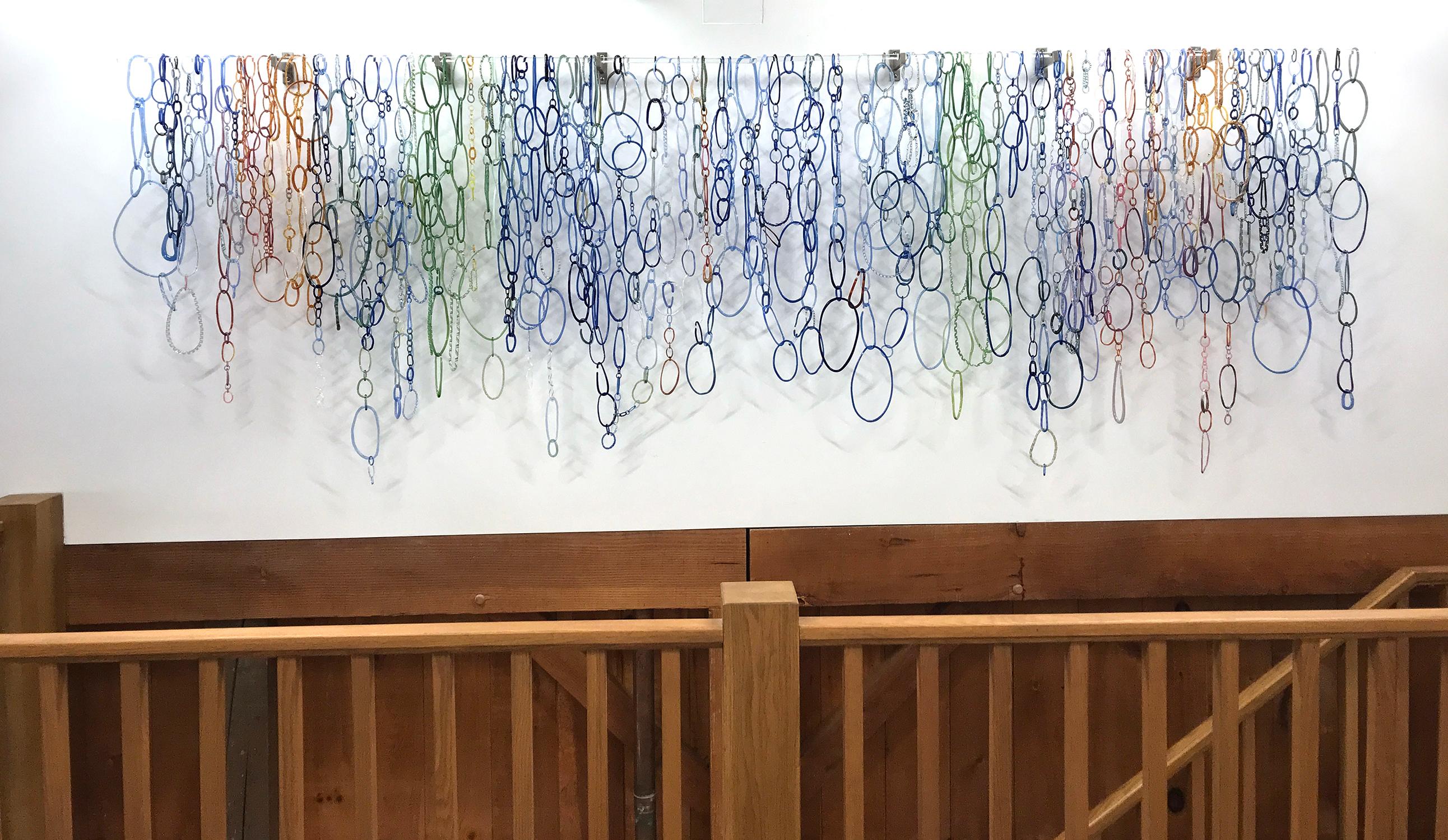 This wide horizontal hanging sculpture is composed of freeform circular loops in various shapes and textures of blue, teal, green, and golden orange with hints of coral pink torch worked borosilicate glass, linked together and hanging from a long