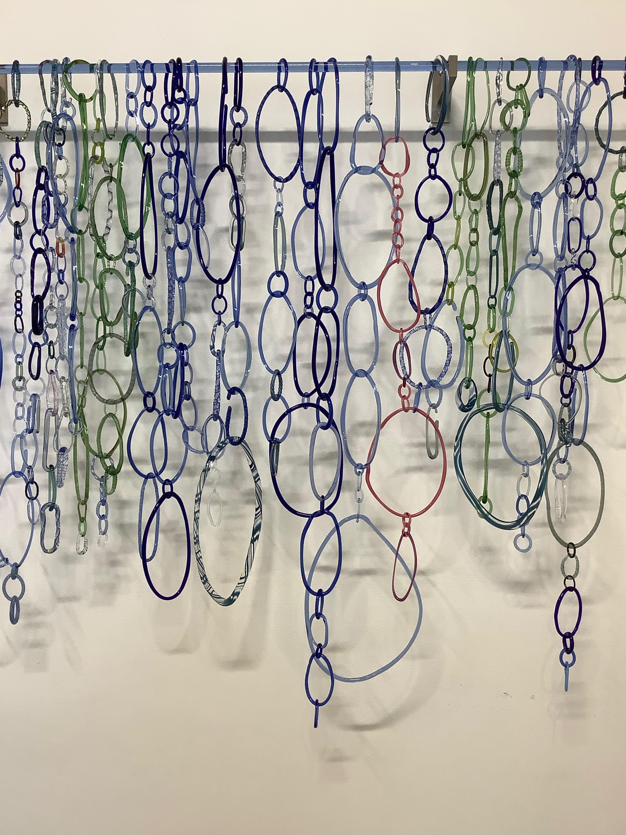 This hanging sculpture by David Licata is composed of free-form circular and oval loops of torch-worked borosilicate glass in dark cobalt blue, light green, light blue, teal and golden pink. The loops, in varying sizes and textures, cascade downward
