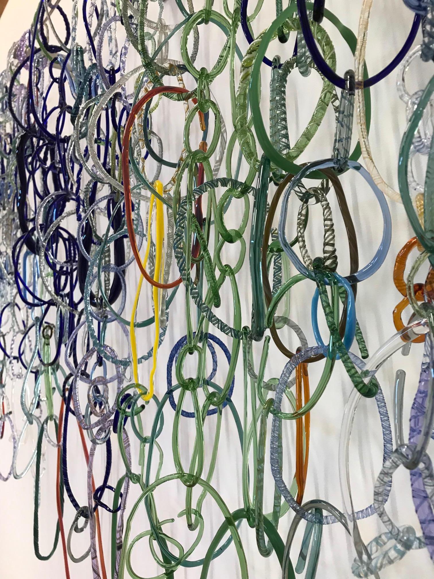 This wide horizontal hanging sculpture is composed of freeform circular loops in various shapes and textures of blue, teal, green, brown and gold torch worked borosilicate glass, linked together and hanging from a long clear glass rod installed with