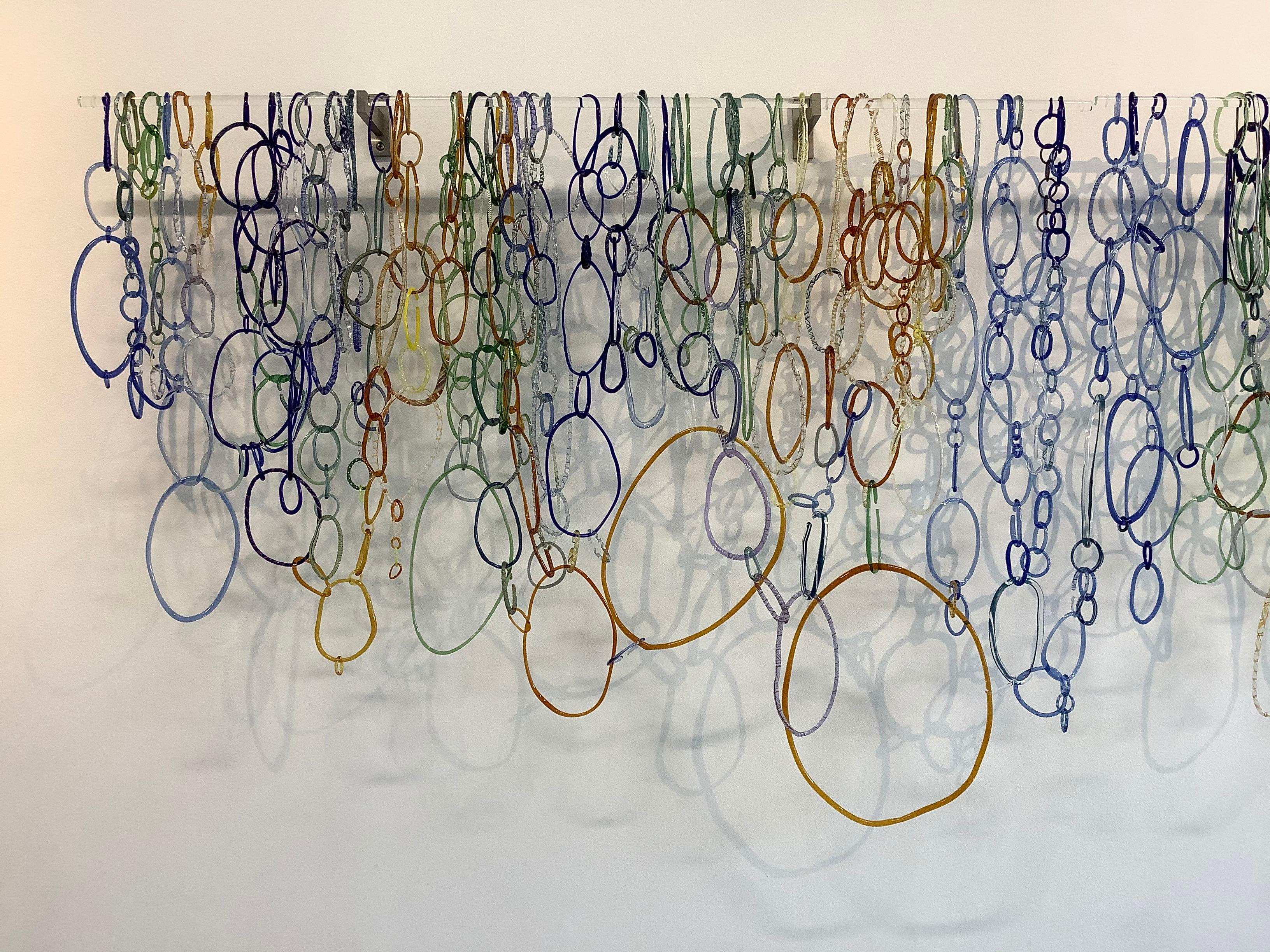 Wide horizontal hanging sculpture composed of freeform circular loops in various shapes and textures of cobalt blue, golden orange, green, pale blue and teal torch worked borosilicate glass, linked together and hanging from two long clear glass rods