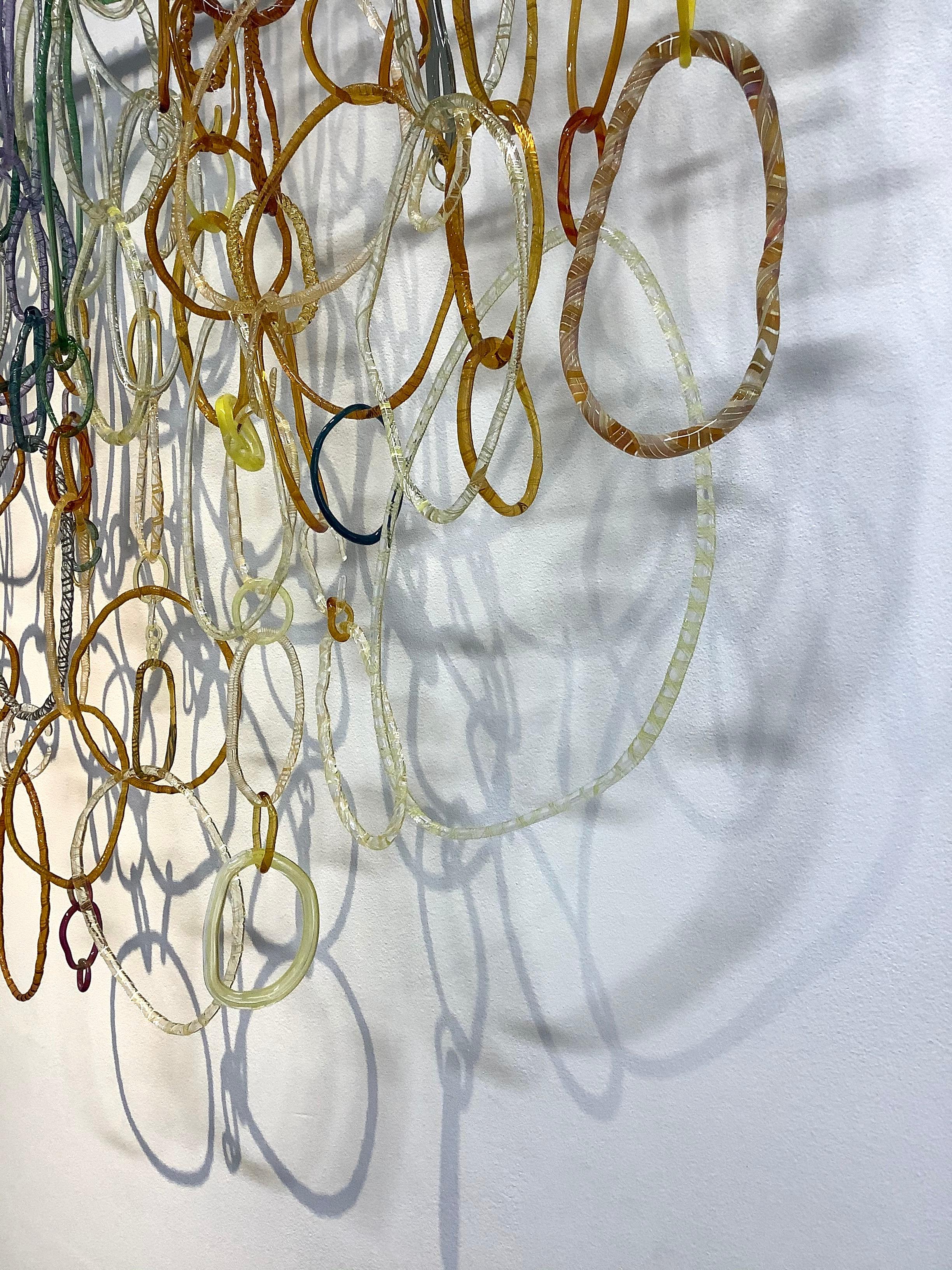 This hanging sculpture by David Licata is composed of free-form circular and oval loops of torch-worked borosilicate glass in golden orange, light green, cobalt blue, pale yellow, and pale lavender. The loops, in varying sizes and textures, cascade