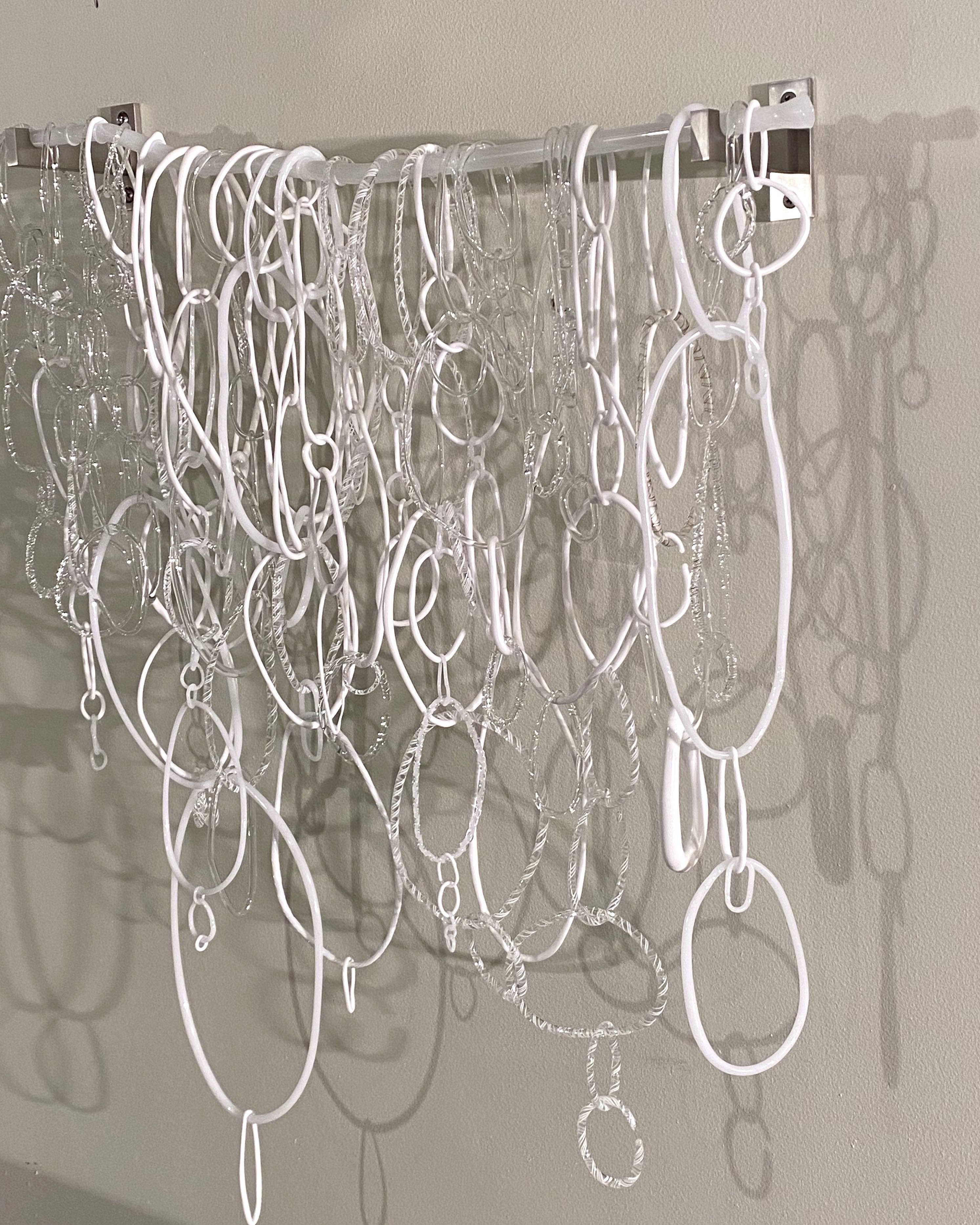 Diese Hängeskulptur besteht aus frei geformten, kreisförmigen Loops in verschiedenen Formen und Texturen aus weißem und klarem, gebranntem Borosilikatglas, die miteinander verbunden sind und an einem unregelmäßig geformten, durchscheinenden weißen